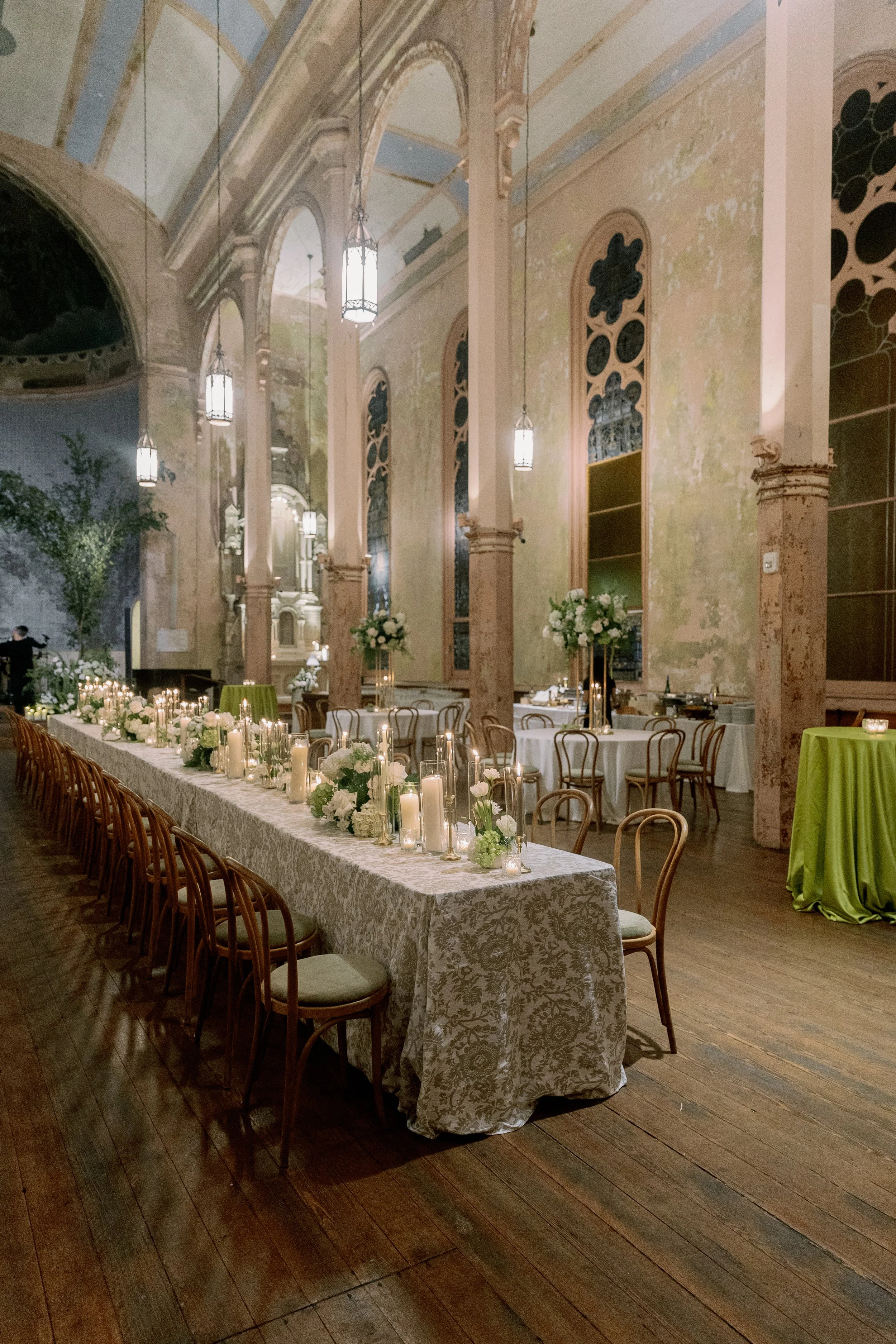 Elegant banquet hall with high arched ceilings, hanging lanterns, distressed pastel walls, tall arched windows, and long decorated tables with candles and floral arrangements, set for a wedding or formal event.