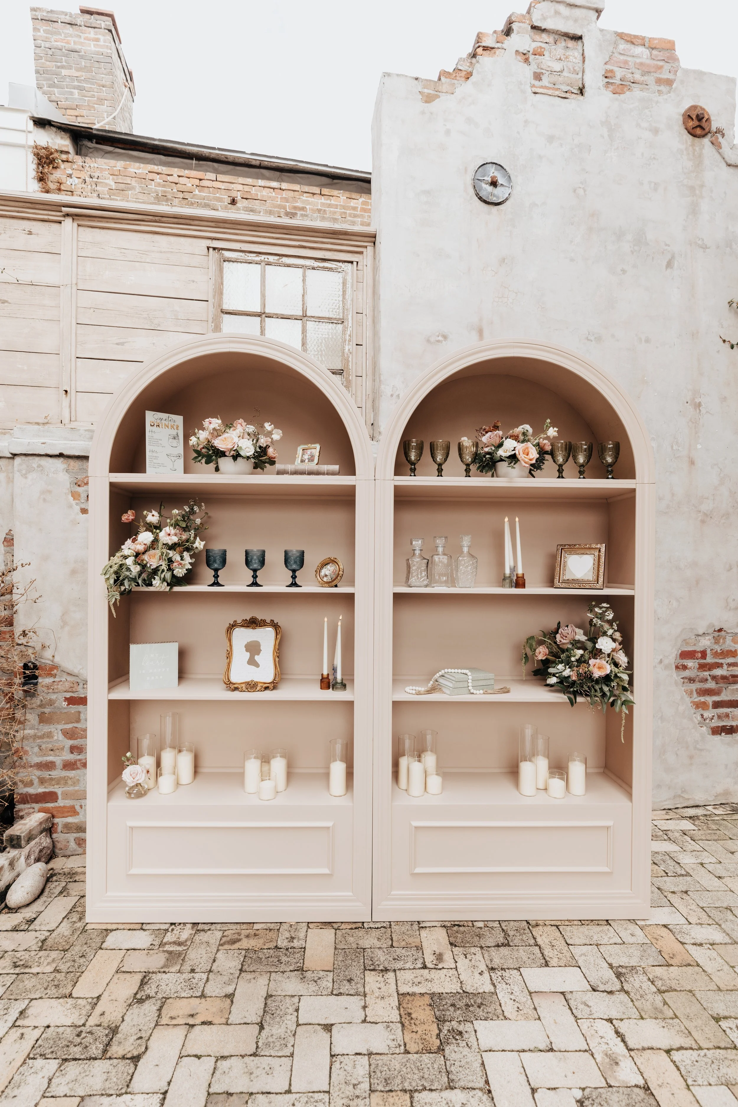 A beige shelving unit with floral arrangements, candles, and decorative glassware against a distressed brick and plaster wall.