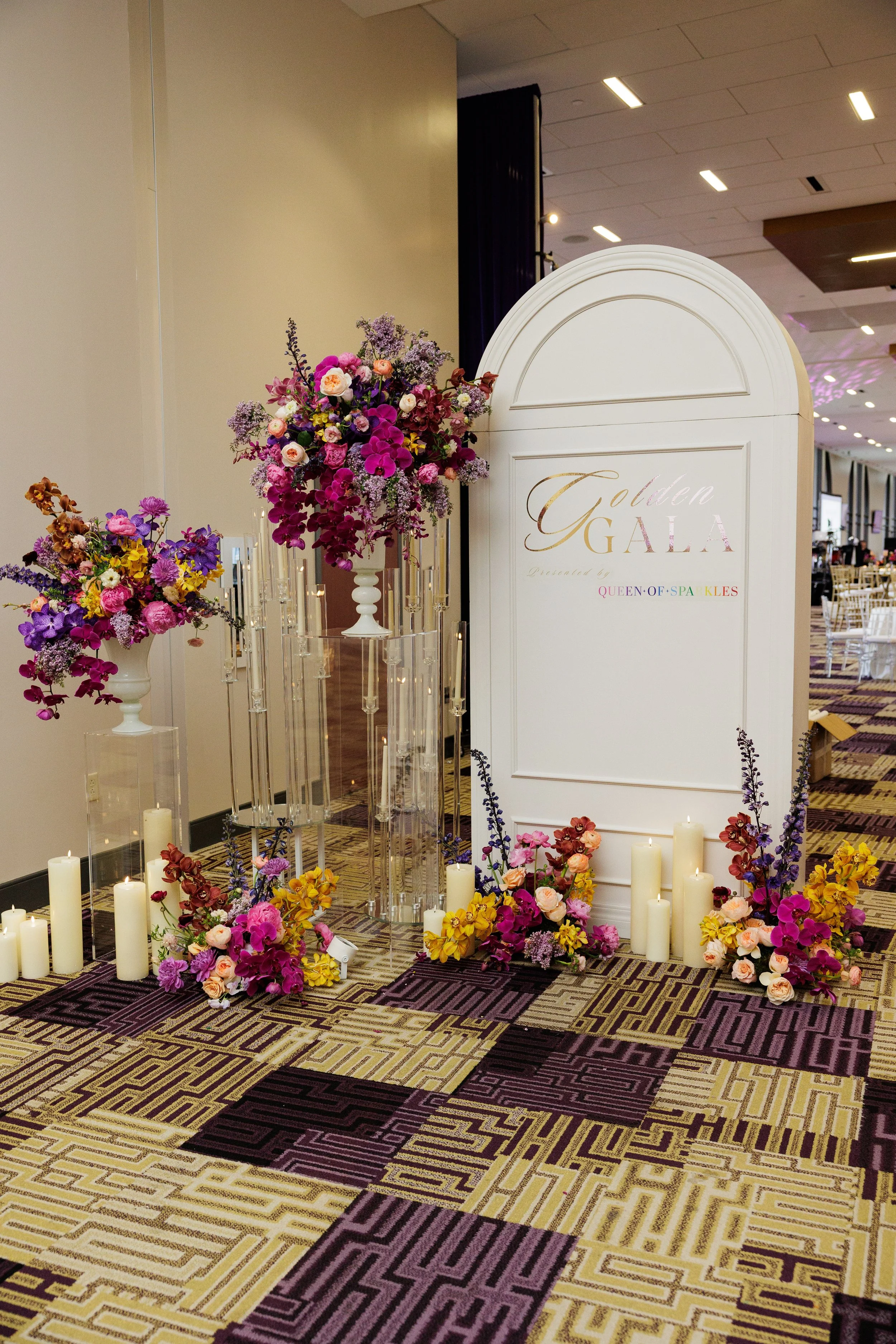 A photo of a floral display and a decorative sign at a formal event or gala. The sign reads 'Golden GALA Present by QUEEN OF SPA KLES' with colorful letters. The floral arrangements include various purple, pink, yellow, and white flowers, with candle