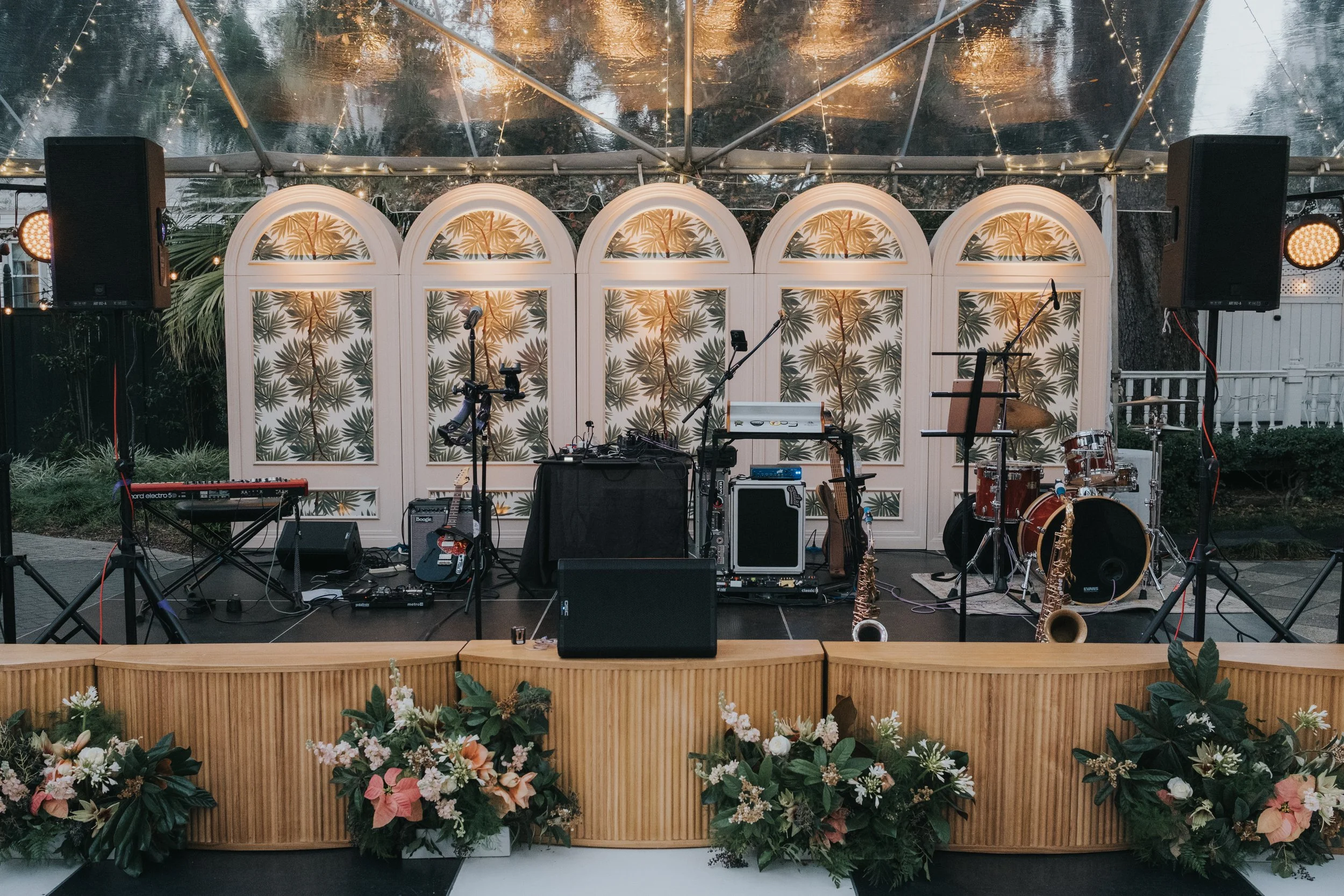 An outdoor stage set up for a live music performance, featuring various musical instruments including drums, saxophones, guitars, and keyboards. The stage has a backdrop with a tropical leaf pattern, surrounded by string lights and two large speakers