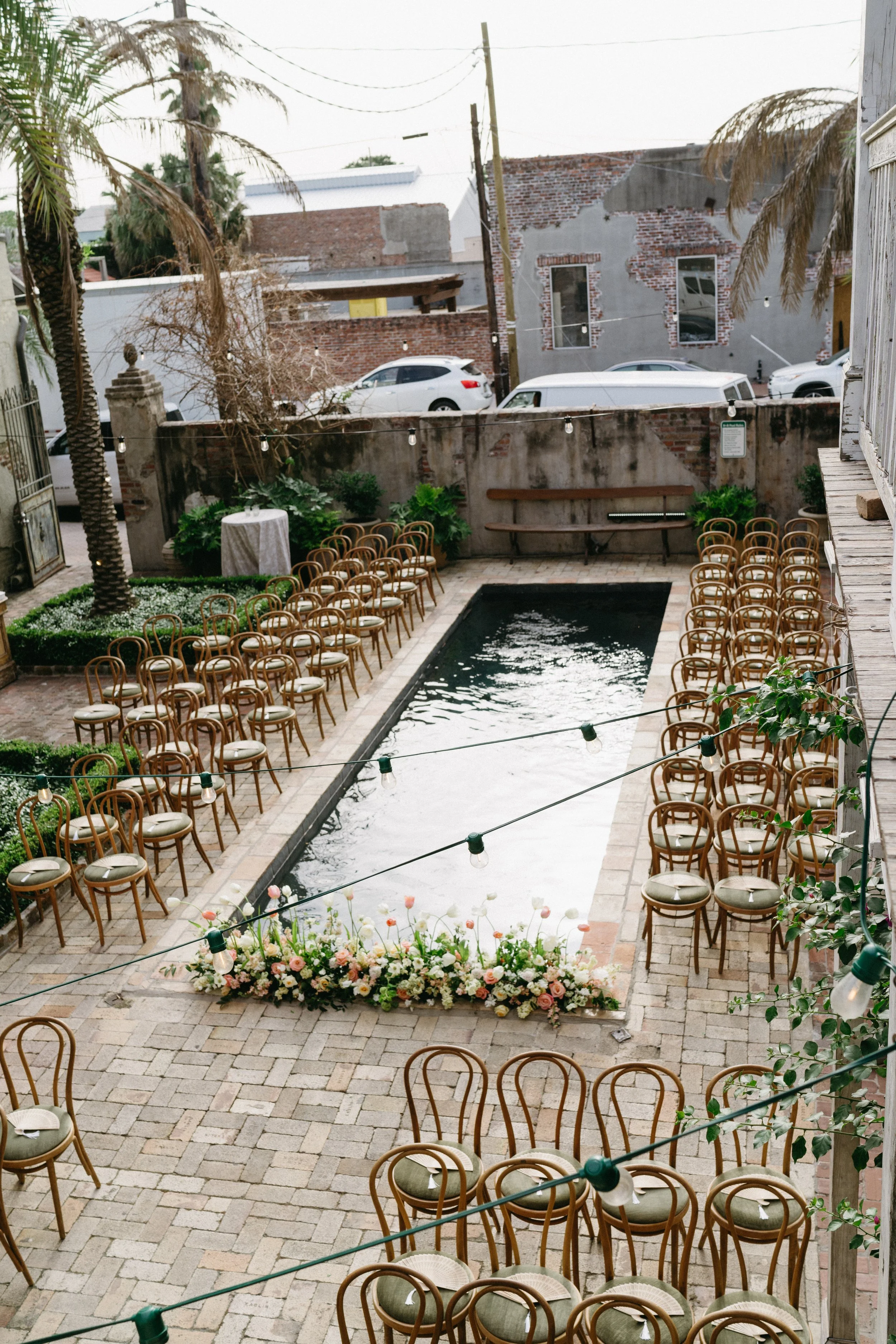 Outdoor event setup with rows of wooden chairs arranged around a small rectangular water feature, decorated with a flower arrangement at one end, string lights hanging overhead, and a brick and concrete wall with buildings in the background.