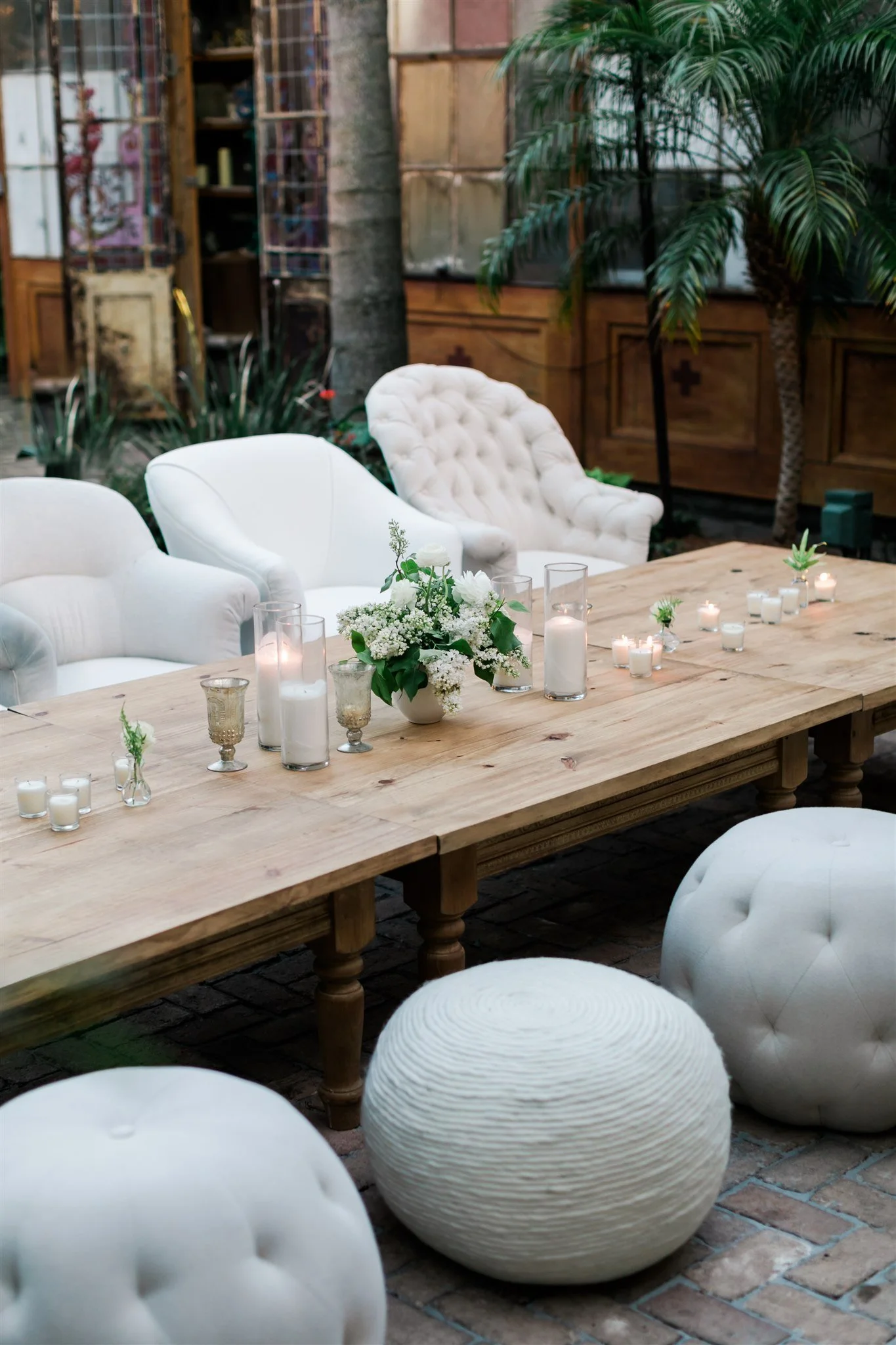 Indoor space decorated with a large wooden table, white tufted upholstered chairs, and white textured poufs. The table has candles, glass vases with white flowers, and small plants. Greenery and indoor plants are in the background.