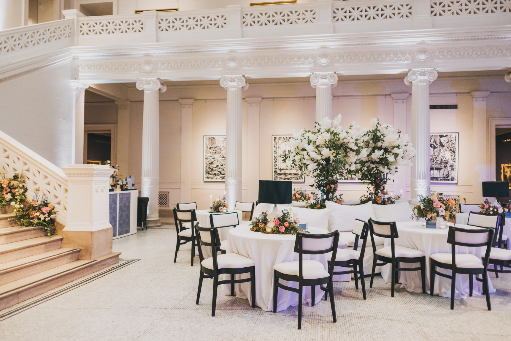 Elegant banquet hall decorated with large floral arrangements, white tablecloths, and black and white chairs, featuring classical columns and framed artwork on the walls.