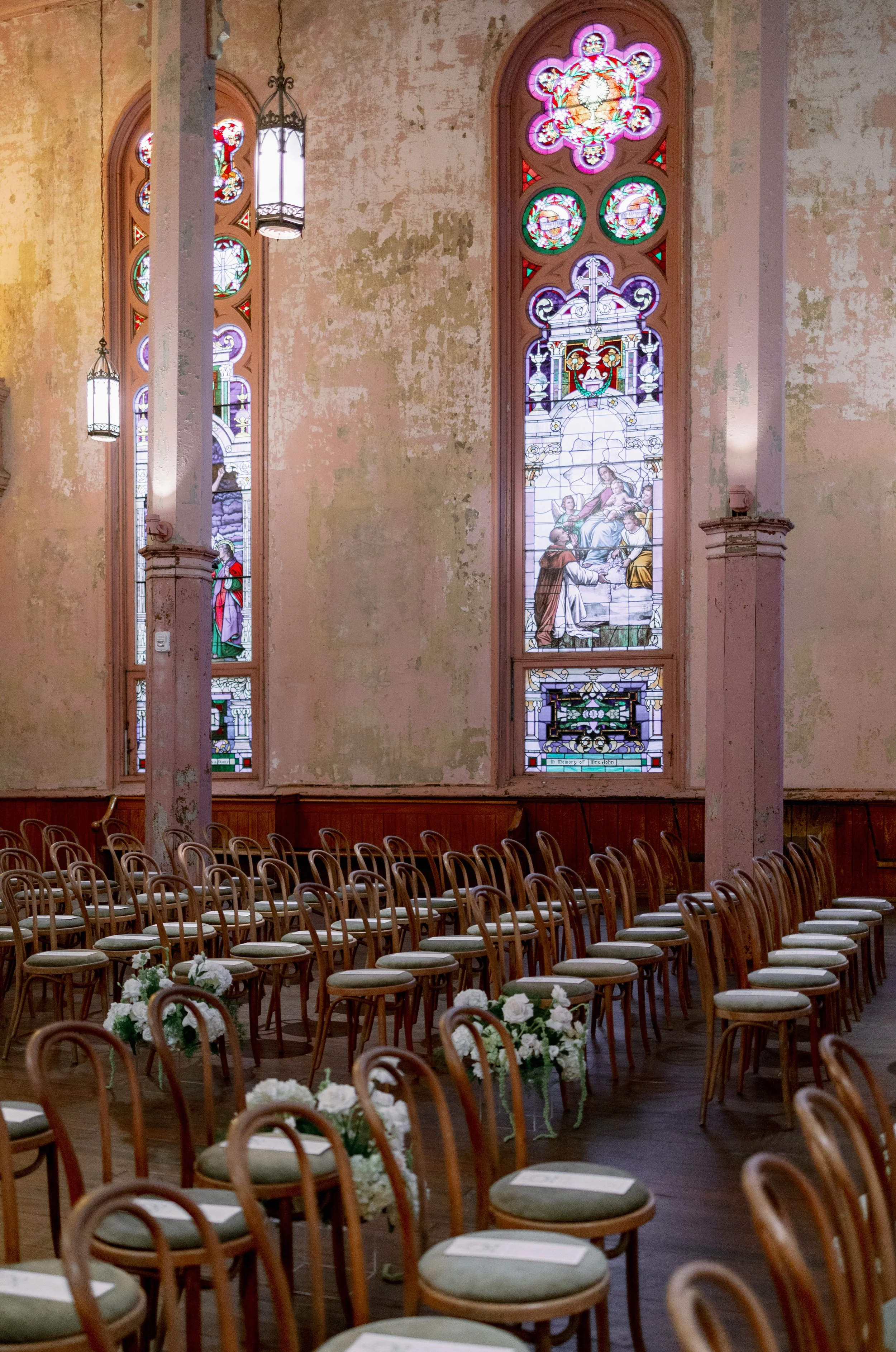 Inside a church with stained glass windows, rows of chairs, and floral arrangements, prepared for a wedding or ceremony.