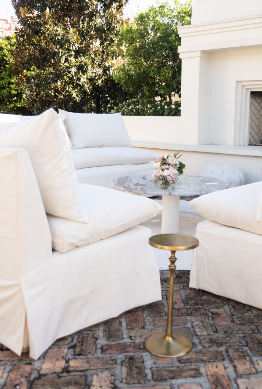 Outdoor patio with white cushioned seating, a small marble table with flowers, a brass side table, and greenery in the background.