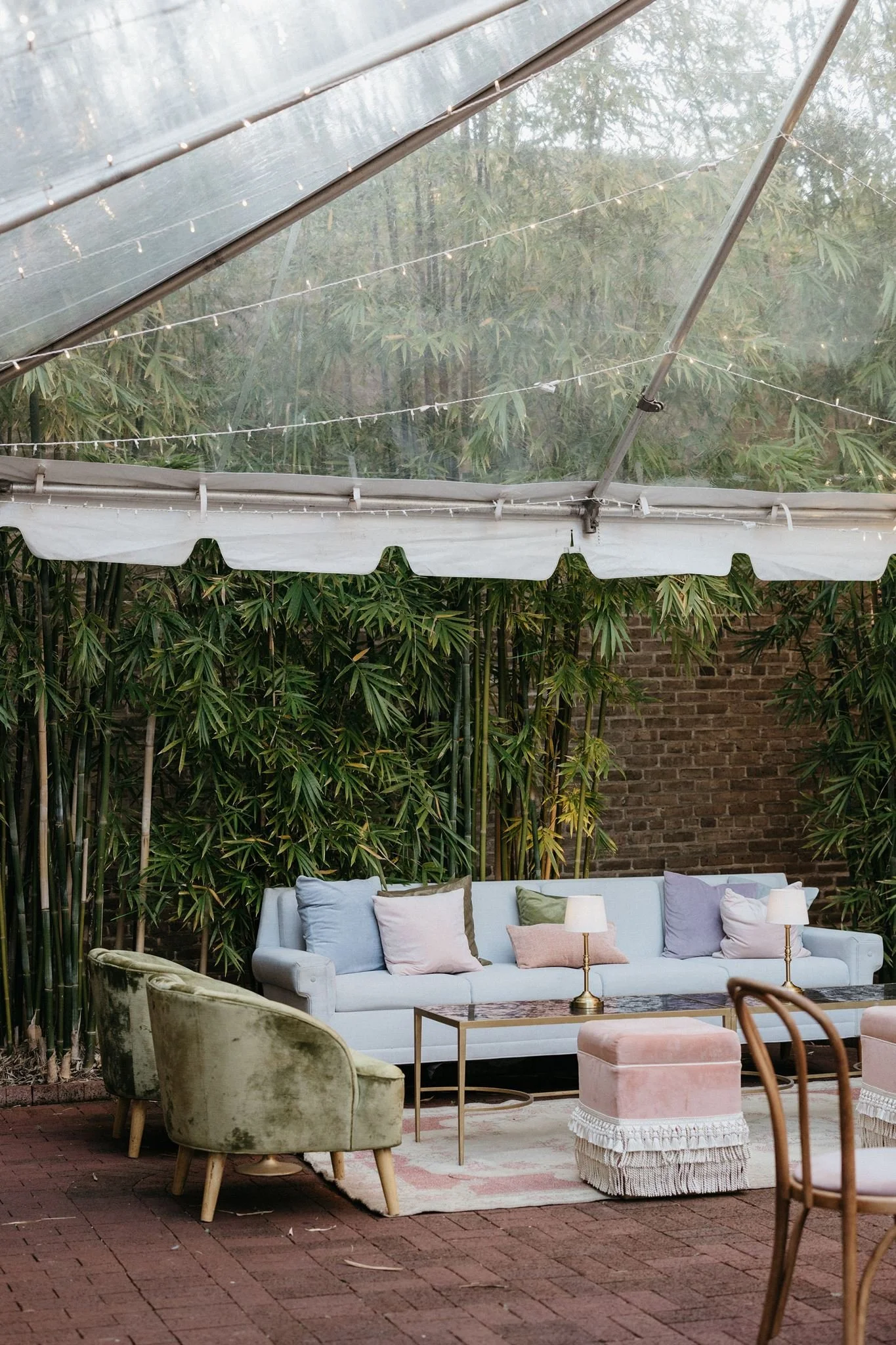 Outdoor patio seating area with a white couch, various colored pillows, small tables, a pink ottoman, vintage velvet armchair, and greenery with a transparent canopy overhead.