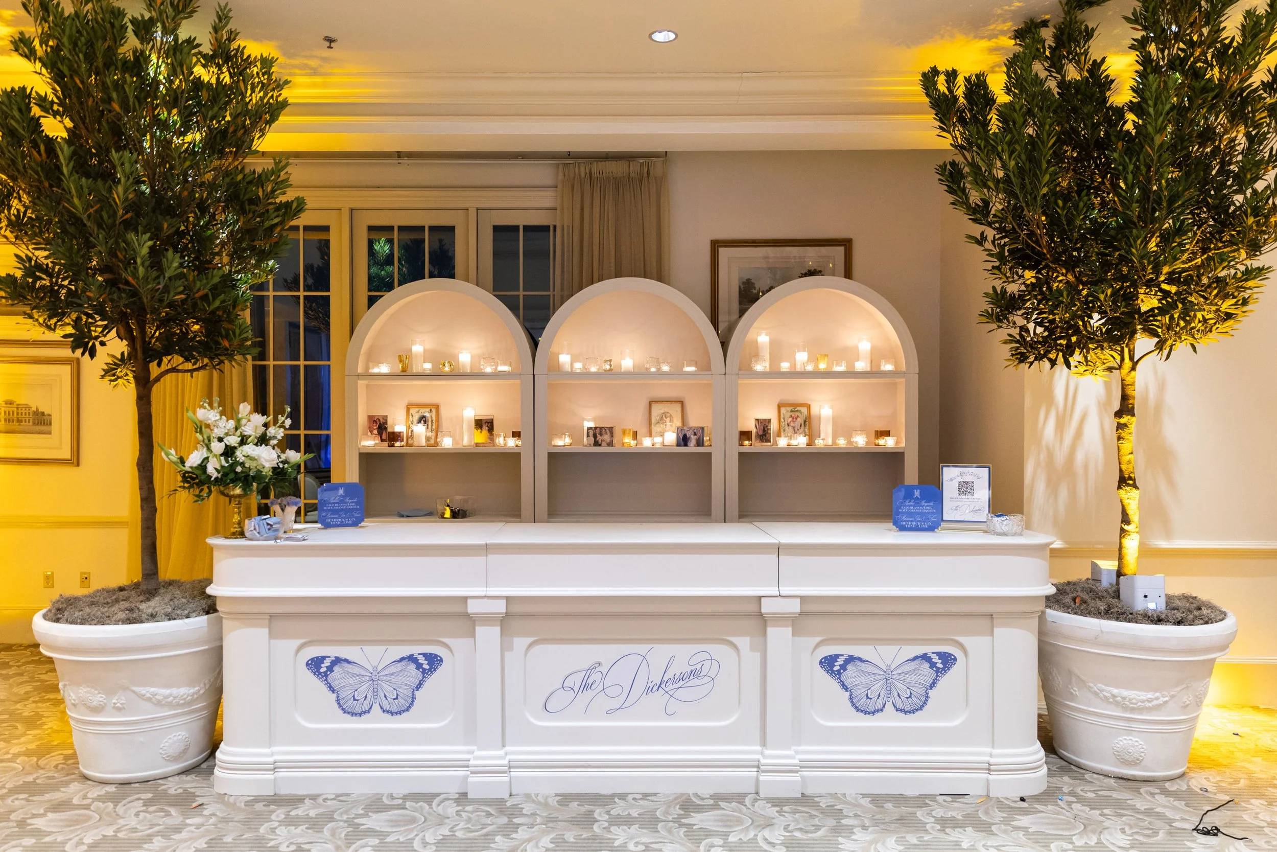 A wedding or event display table with three white arched shelves filled with candles and framed photos, flanked by large potted trees, in a decorated indoor venue.