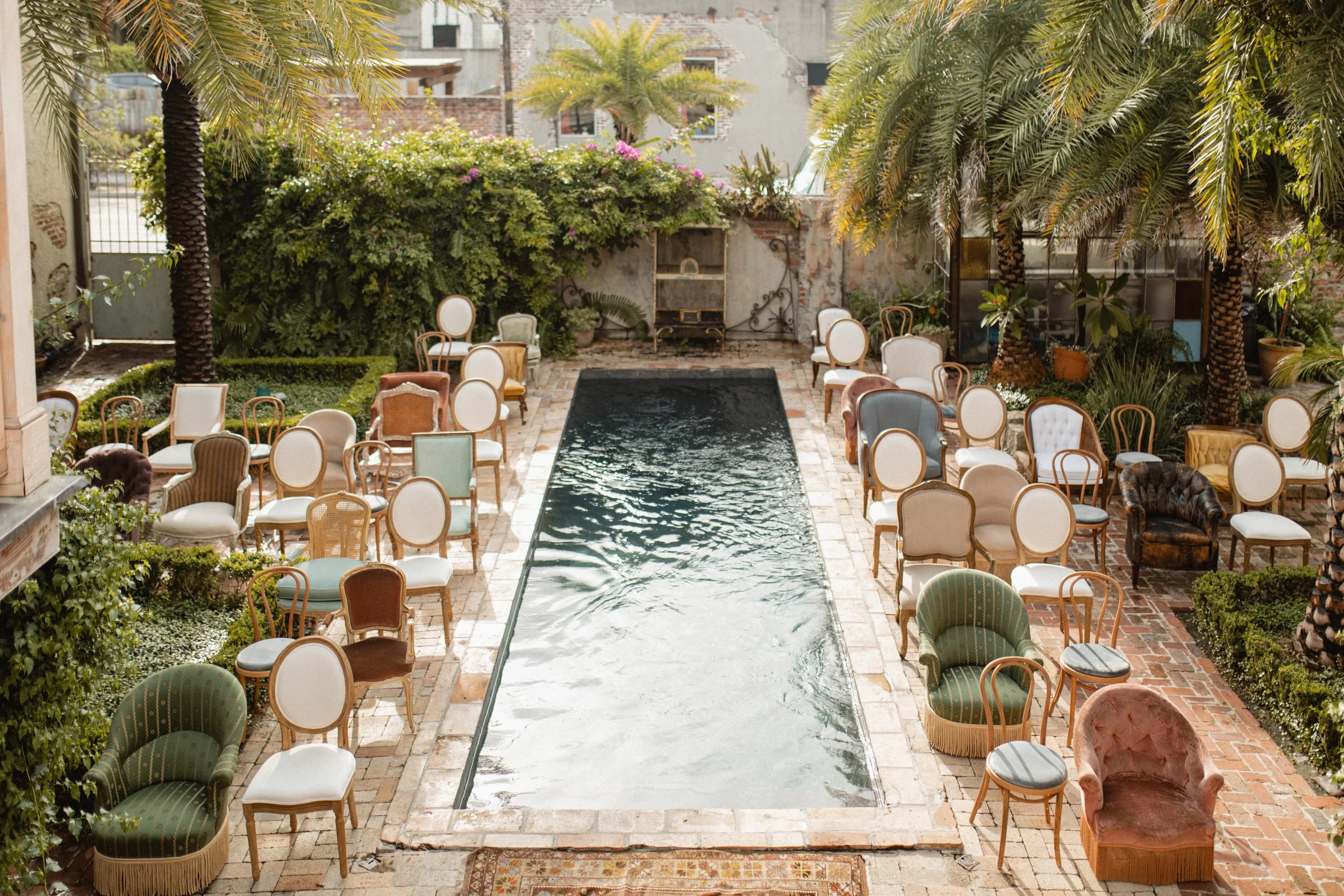 An outdoor patio with a rectangular water feature in the center, surrounded by numerous vintage-style chairs and sofas with various upholstery, set on brick paving. There is lush green foliage and palm trees along the perimeter, with a weathered wall