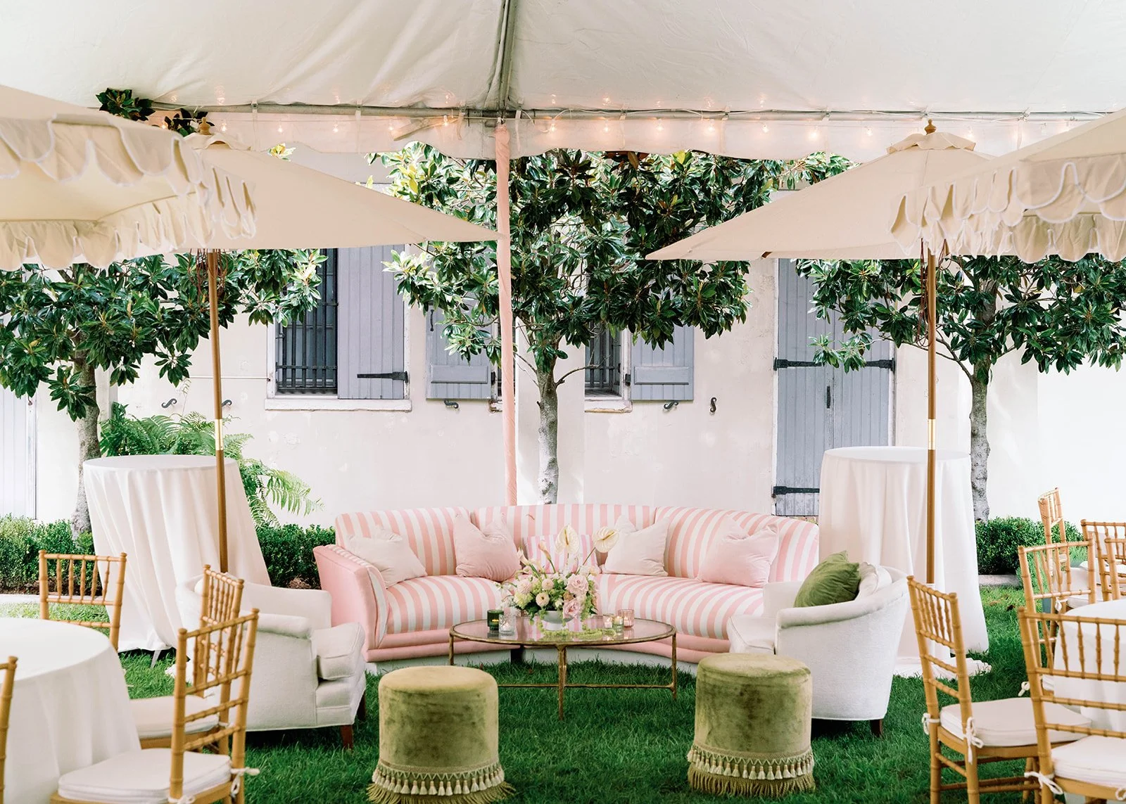 Outdoor patio with a pink and white striped sofa, surrounded by white and beige chairs, green ottomans, and large umbrellas. Green trees and a white building with gray shutters are in the background.