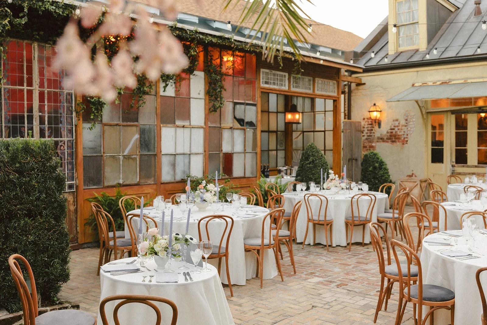 Elegant outdoor dining setup with round tables covered in white tablecloths, floral centerpieces, candles, glassware, and silverware, surrounded by wooden chairs, with a rustic building and string lights in the background.