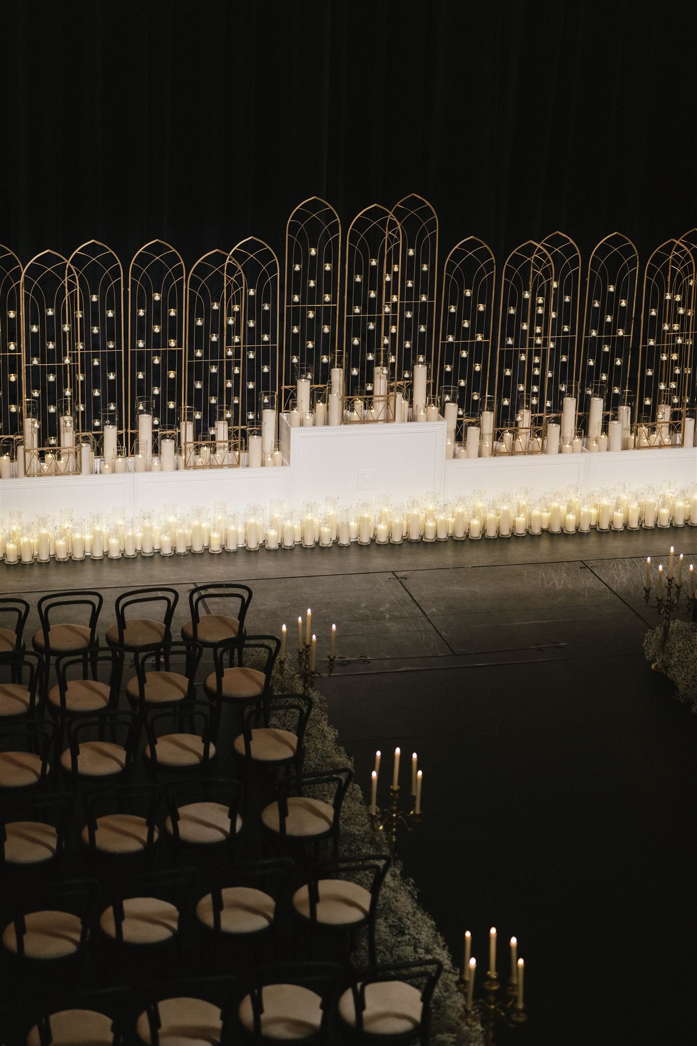 Ceremonial stage decorated with numerous white candles and gold-colored gothic-style arched framework with small hanging lights against a dark backdrop.