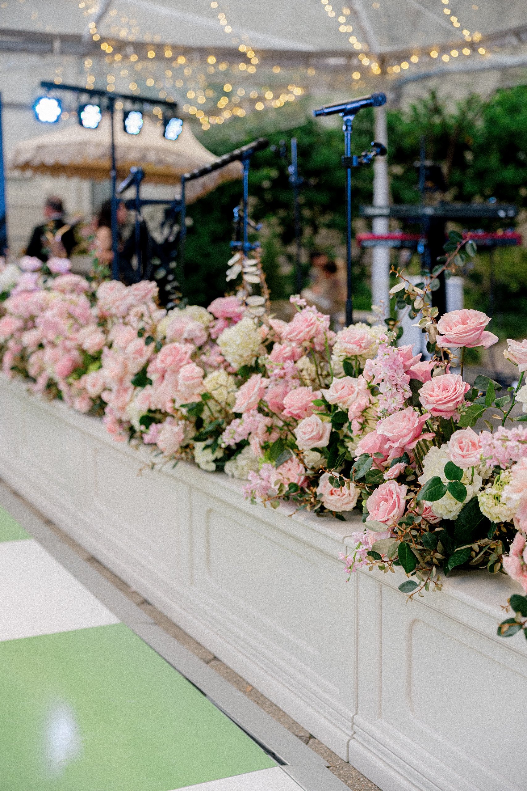 A white display table decorated with pink and white flowers, with microphones and music stand in the background under string lights at an outdoor event.
