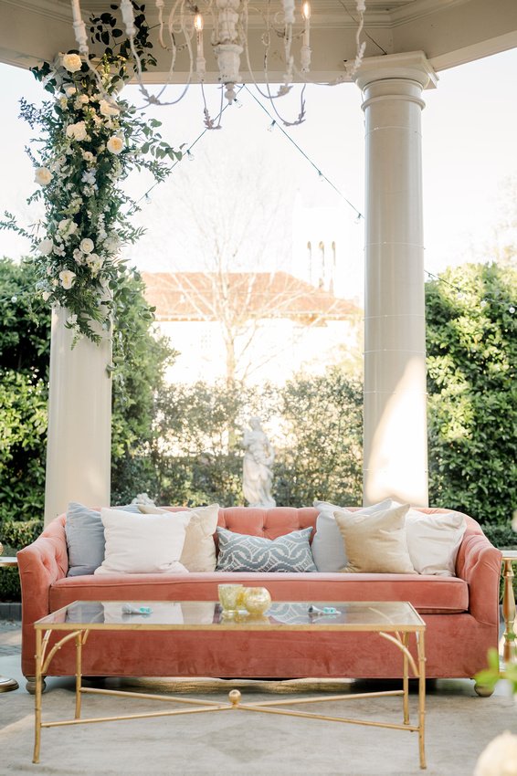 A cozy outdoor seating area with a pink velvet sofa and several throw pillows, a glass-top coffee table, and a white chandelier hanging above, set against a lush green background with trees and a statue.