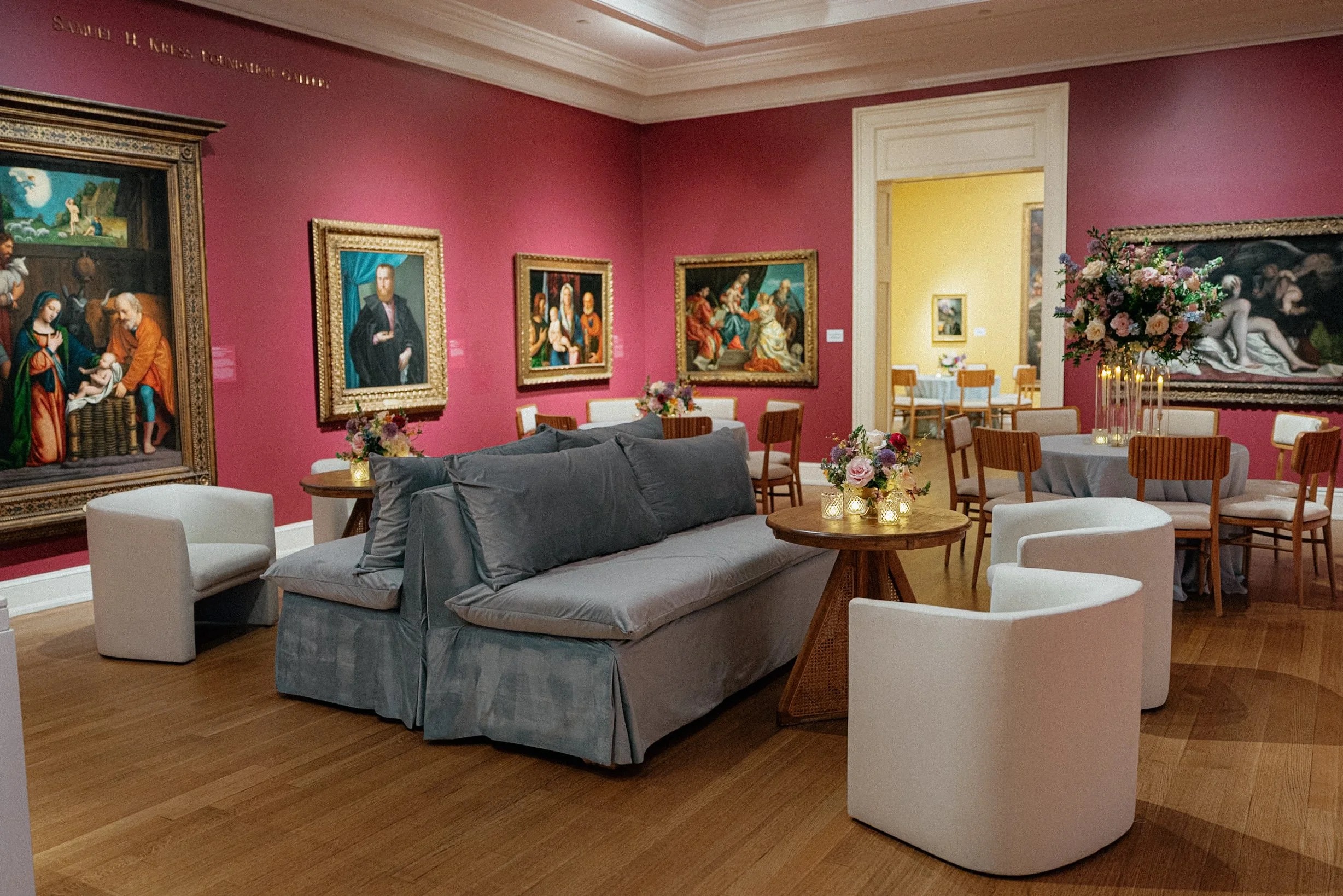 An art gallery with pink and yellow walls, featuring classical paintings, modern furniture, and floral arrangements with candles.