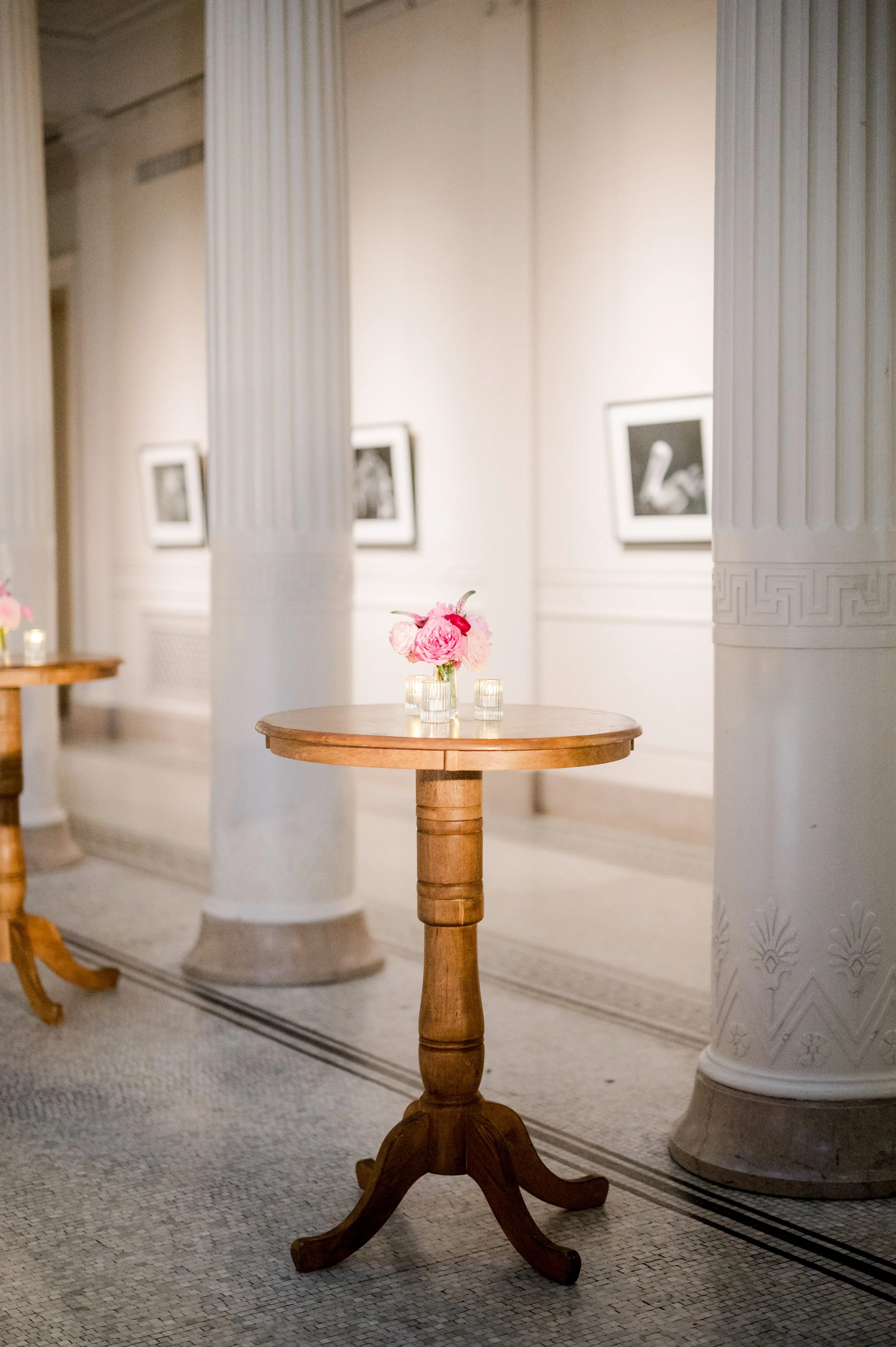 A round wooden table with a flower arrangement in a glass vase and candles, situated in a large room with ornate columns, framed photographs on the wall, and patterned tile flooring.