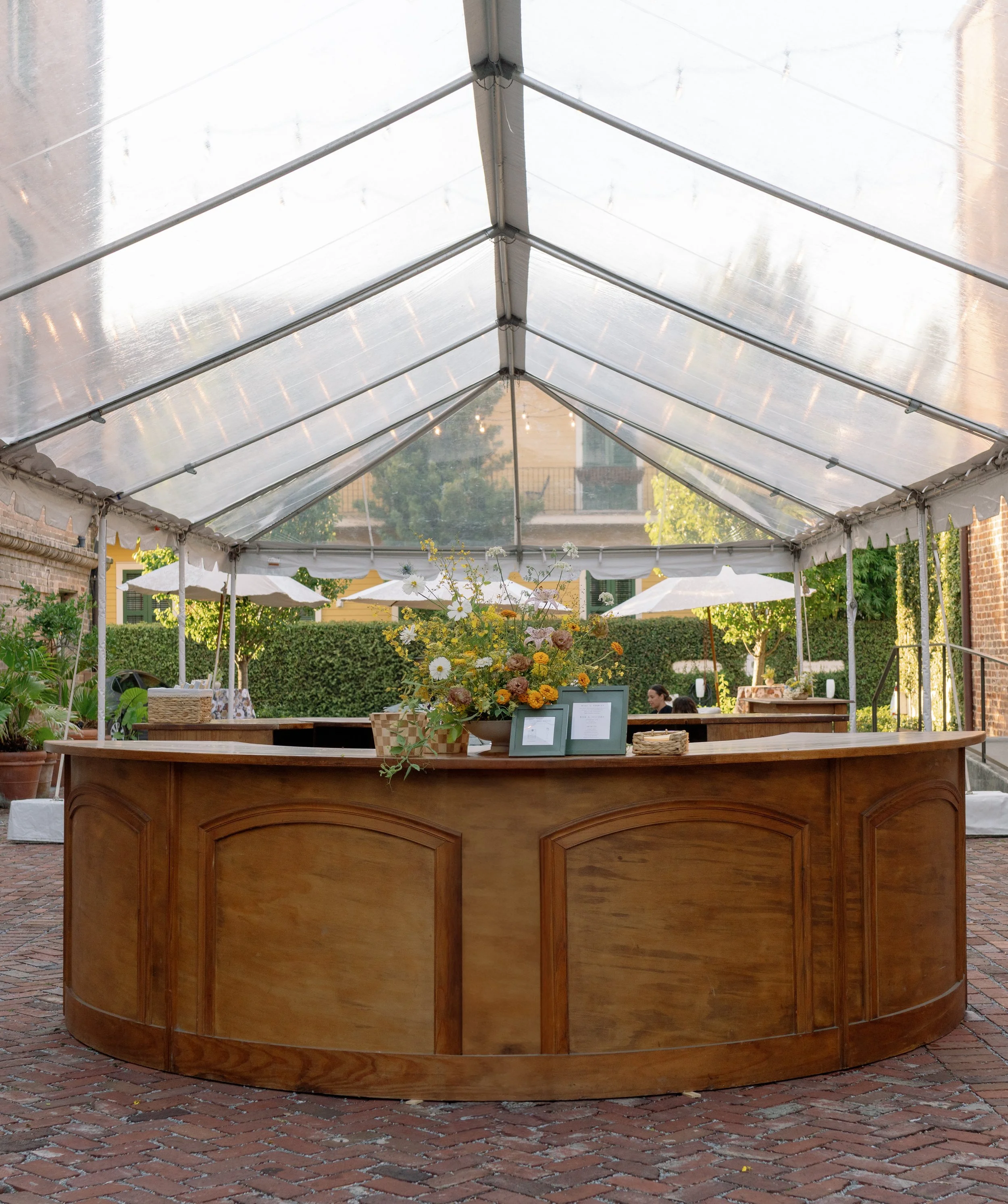 Outdoor event space with a tent and a wooden bar or counter, decorated with a vase of flowers, set on brick pavement with some greenery in the background.