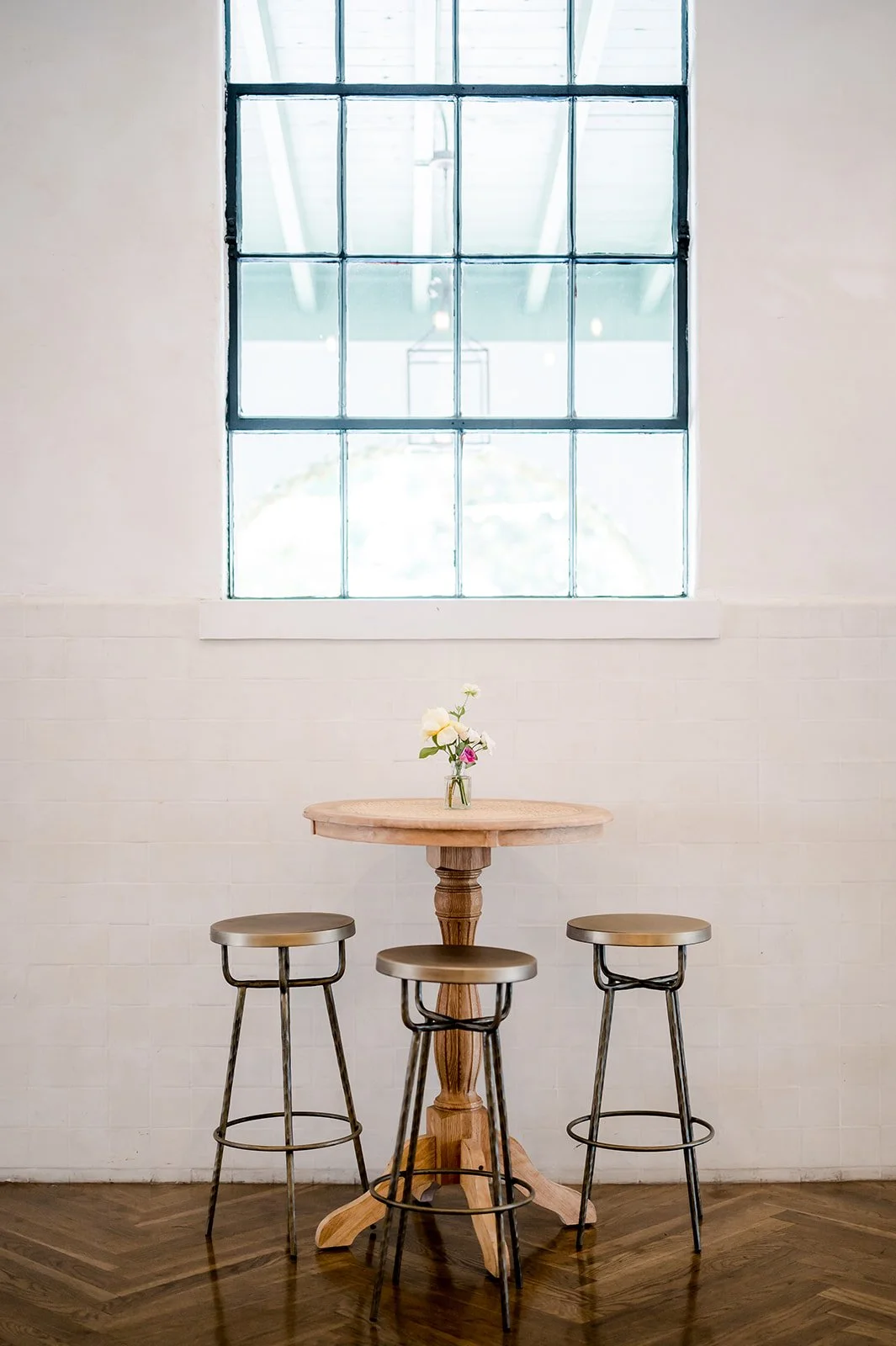 A wooden table with a small glass vase holding white and pink flowers sits beneath a large industrial-style window on a brick wall. Three metal barstools surround the table, and the floor is wood.