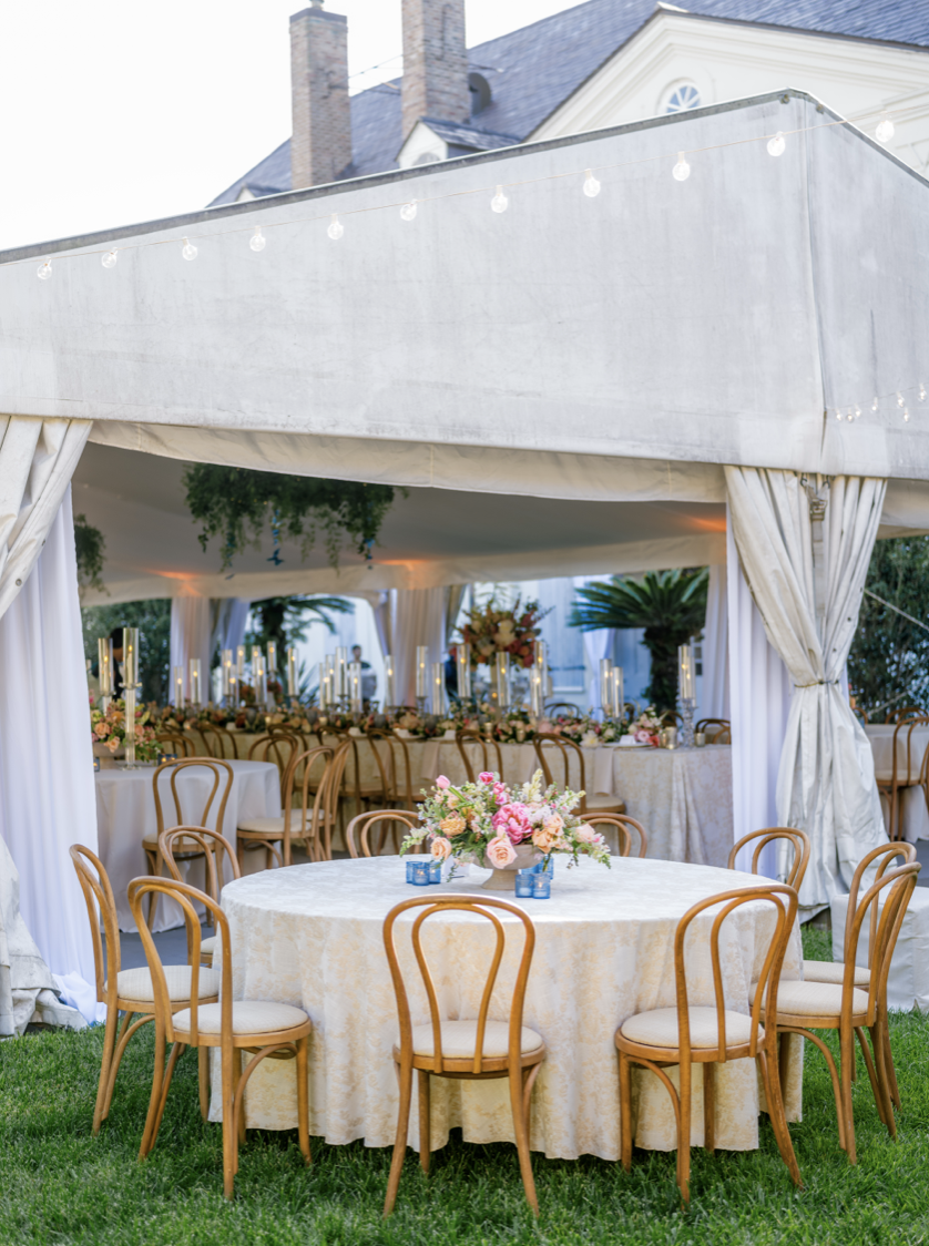 Wedding reception setup with round table, pink and white floral centerpiece, and chairs outdoors under a white tent with string lights.