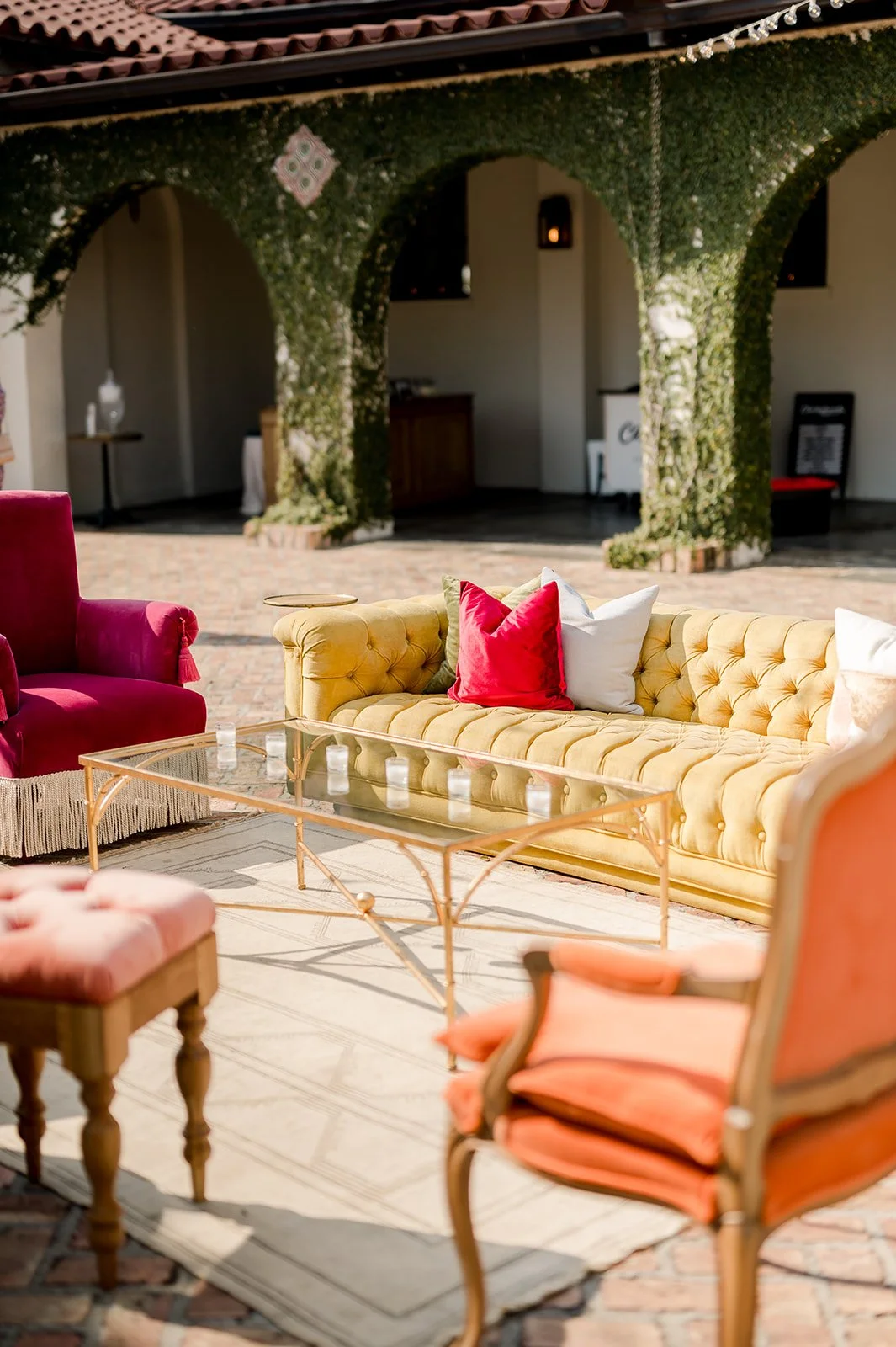 Outdoor patio with a yellow tufted sofa adorned with red, white, and green pillows, a pink armchair, and a glass coffee table on a beige rug, with a background of a building with arches covered in greenery.