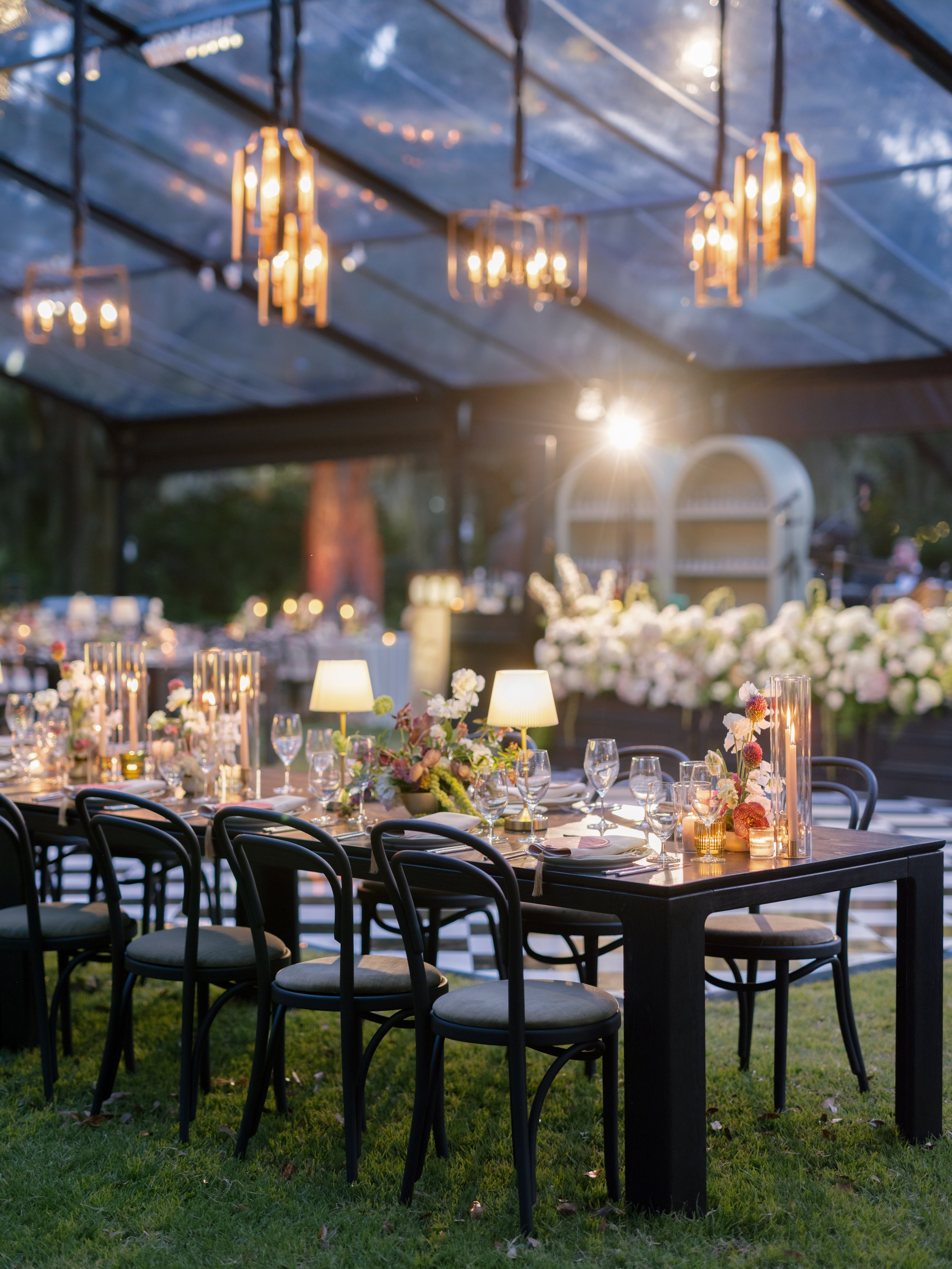 Elegant outdoor dining setup with a black table, surrounded by black chairs, decorated with flowers, candles, and lamps, under a canopy with hanging lights, during evening time.