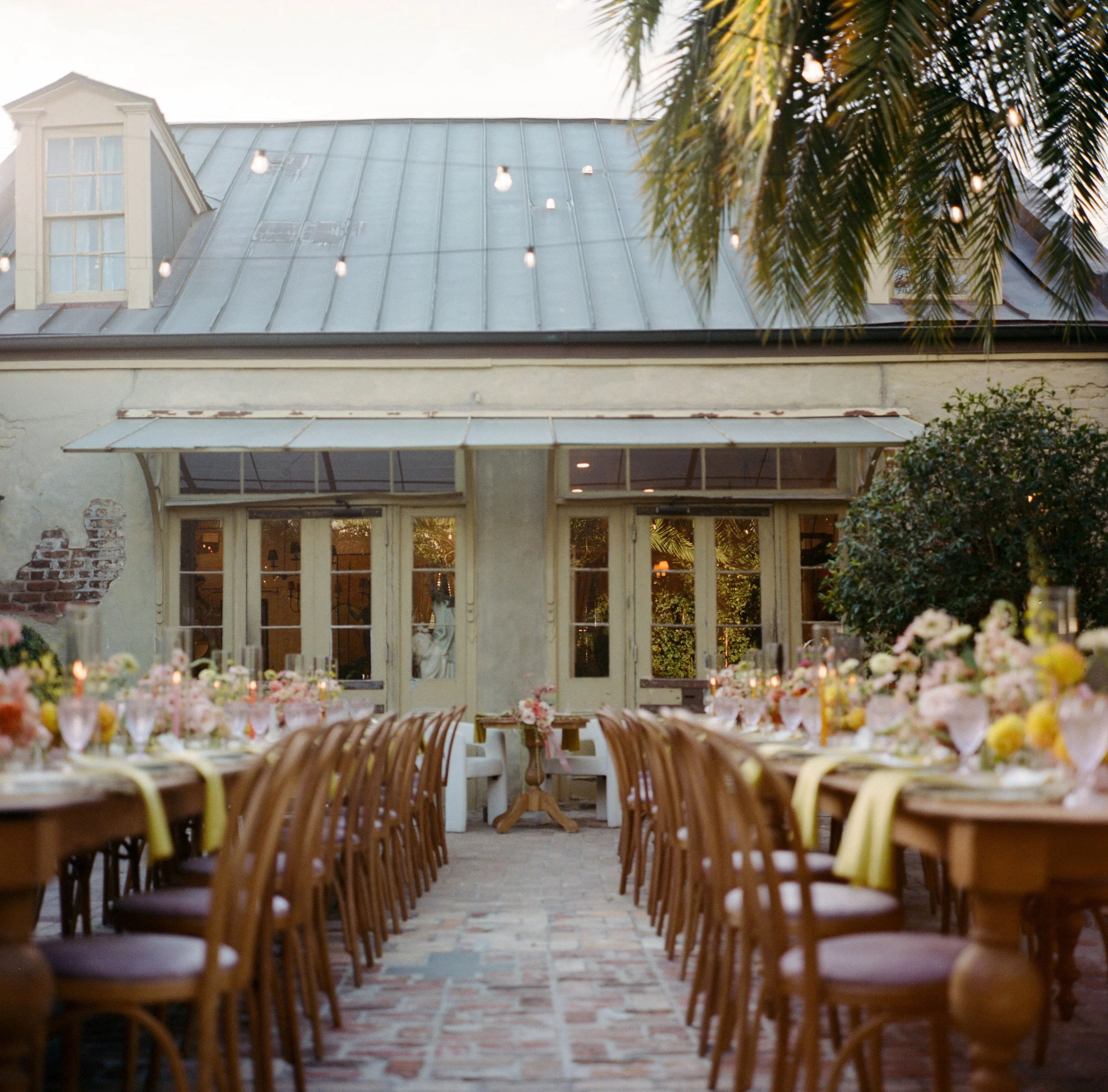 Indoor dining area decorated for an event with round tables, chairs, and floral centerpieces, with a large windowed wall and string lights overhead.