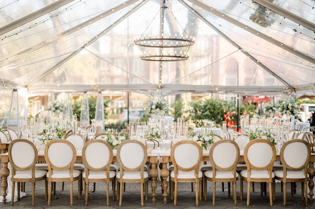Wedding reception setup with a long table decorated with white flowers, glass candle holders, and white chairs, under a clear tent with string lights and a chandelier.