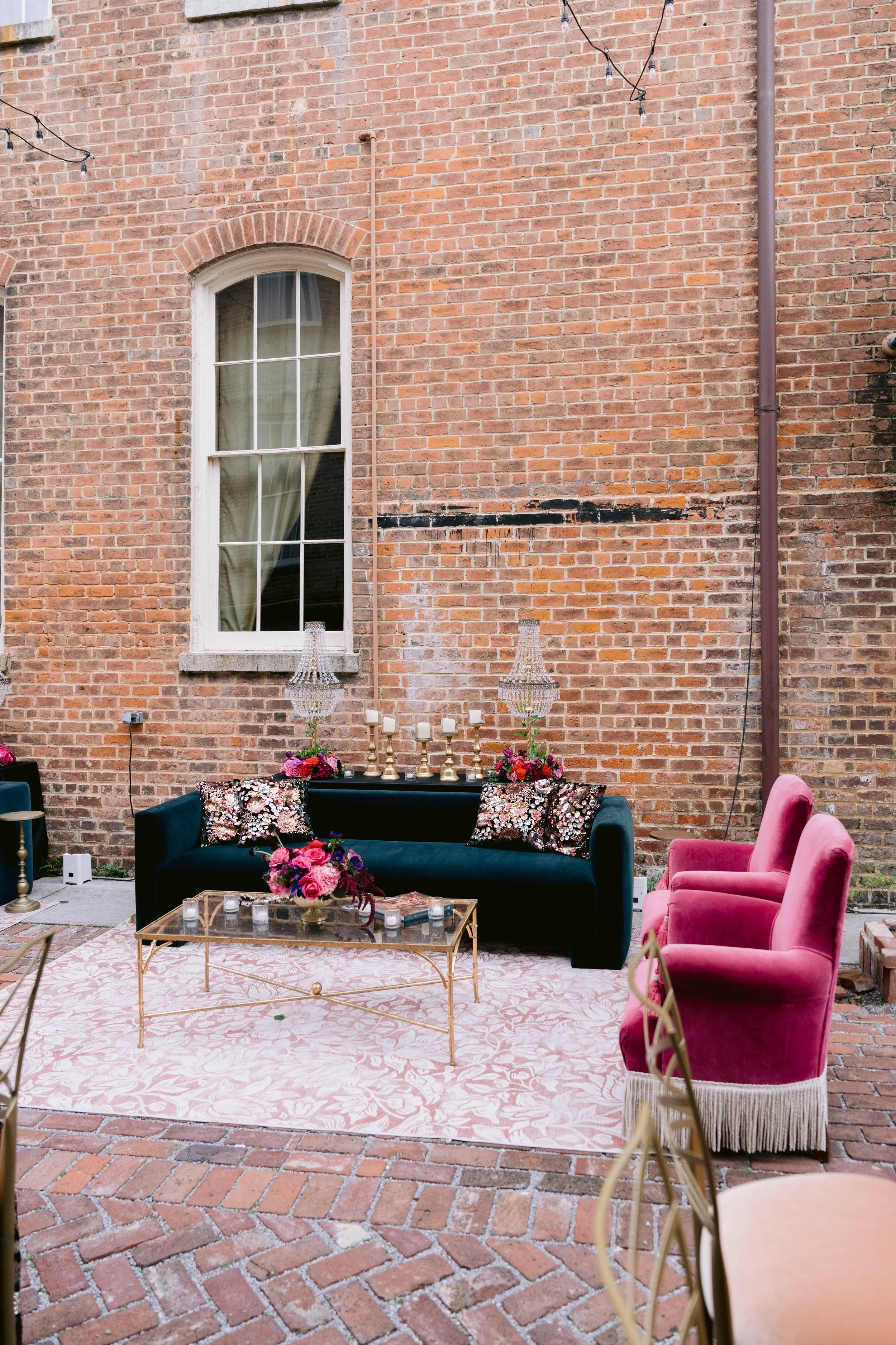 Outdoor patio decorated for an event with pink and black seating, floral arrangements, candles, and string lights against a red brick wall.