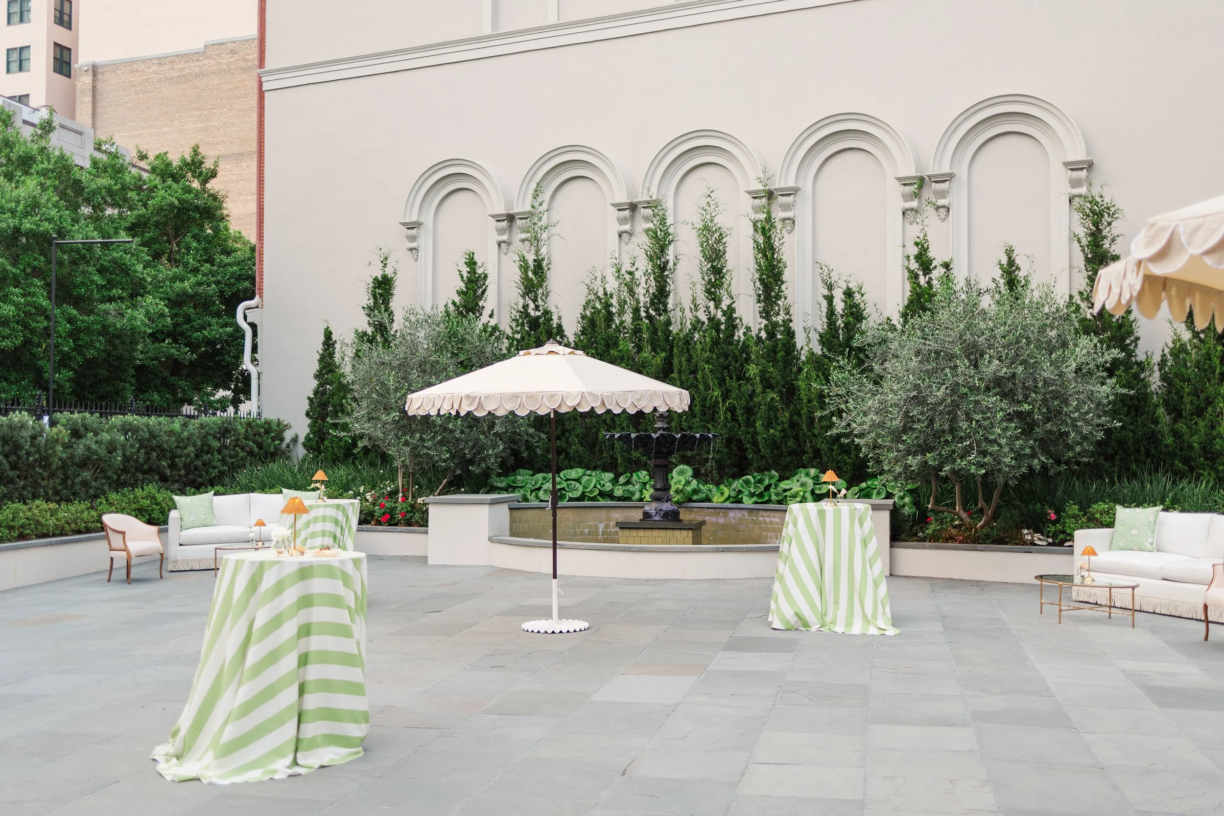 An outdoor patio area with tables covered in green and white striped tablecloths, umbrellas, plush white seating with green pillows, lush green bushes, and a decorative fountain in front of a white building with arched architectural details.
