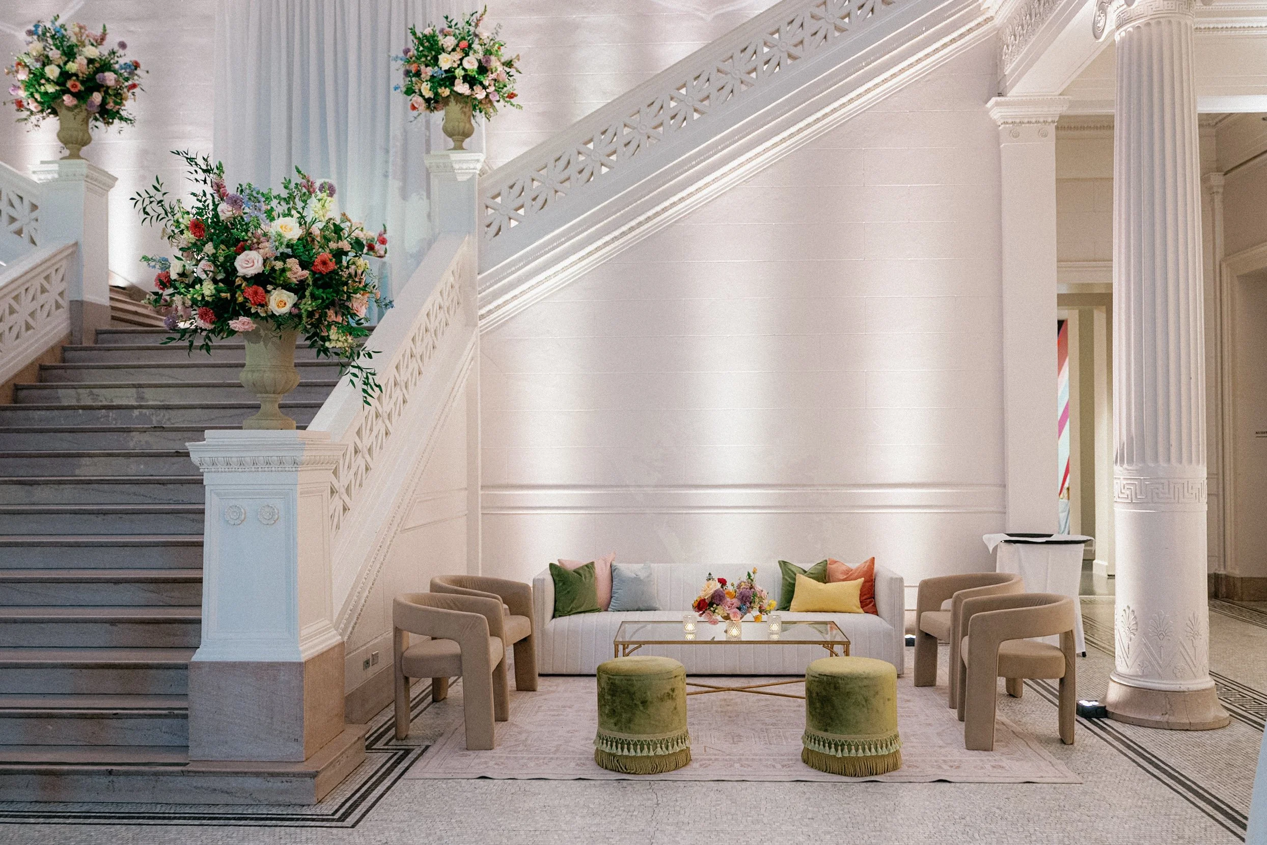 Interior of a grand, elegant hall with white walls, a staircase decorated with flowers, surrounded by classical columns and ornate moldings, with a sitting area featuring a white couch, colorful cushions, and small green stools.
