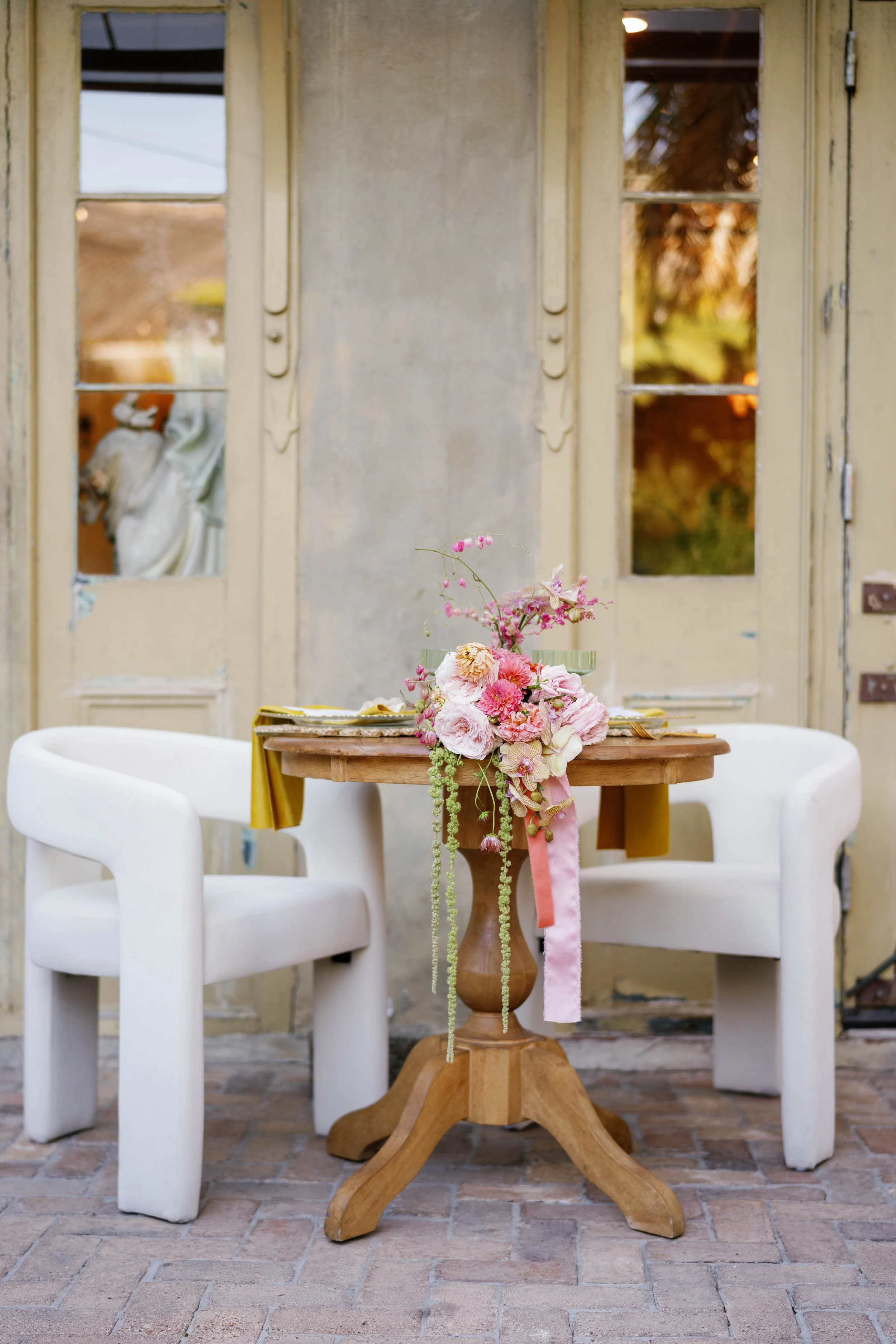 A wooden table with a floral centerpiece, surrounded by two white chairs, set in a rustic outdoor patio with brick flooring and a weathered yellow door in the background.