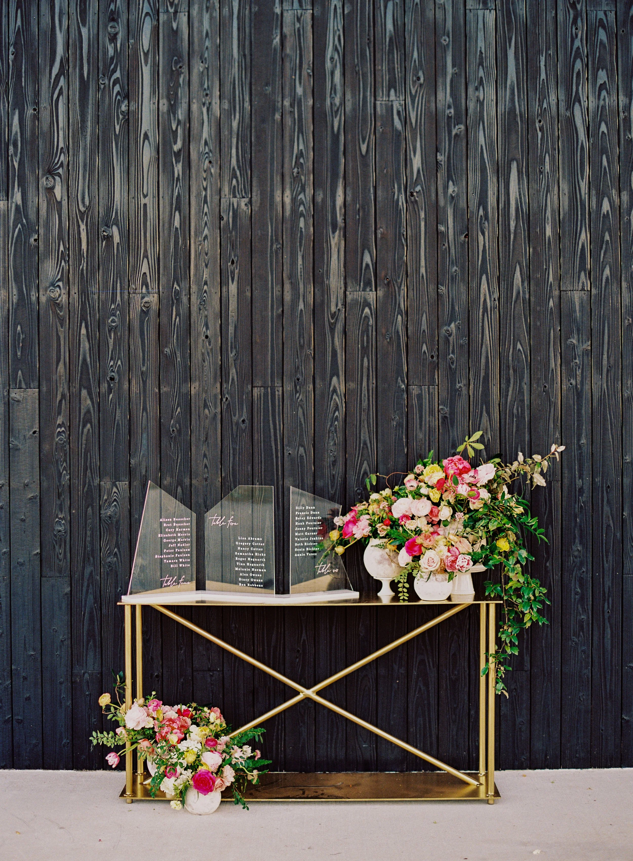A black wooden wall with a table in front of it, holding a clear acrylic memorial display with names of people. Next to the display, there is a large white vase with pink and white flowers, and a small white bouquet to the side.