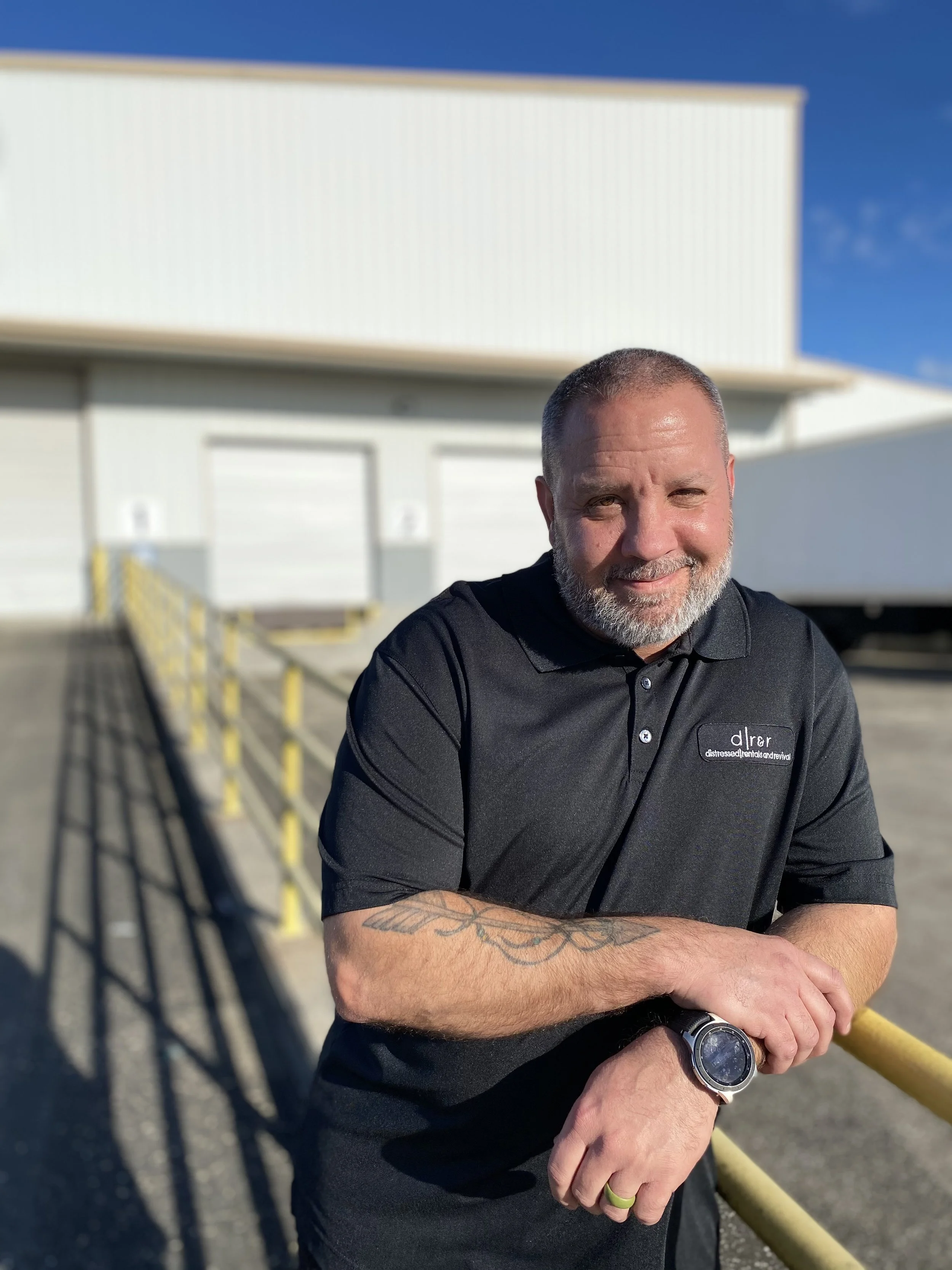 A man with a beard and short hair standing outdoors, leaning on a yellow railing, wearing a black polo shirt with a logo that reads 'dler stress additive and removal', a watch on his left wrist, and a wedding band on his left ring finger. The background features a white industrial building and a clear blue sky.