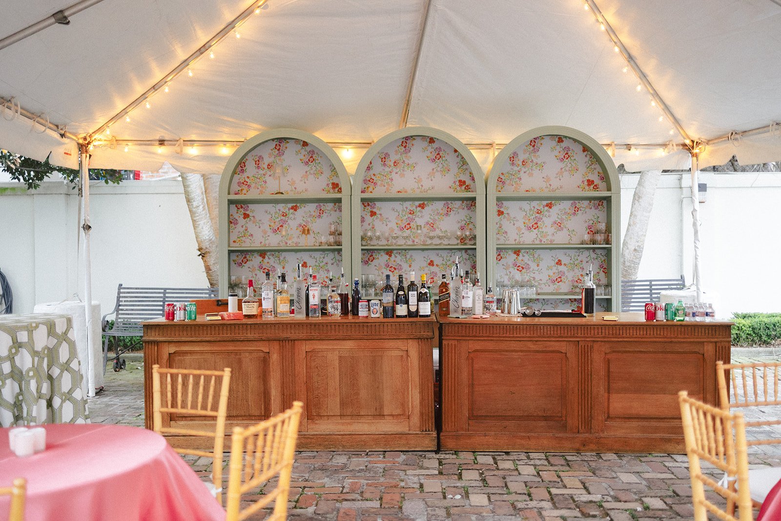 Outdoor bar setup under a white tent with string lights, featuring a wooden counter with bottles of alcohol, mixers, and drinkware, and patterned wallpaper-backed shelves behind.
