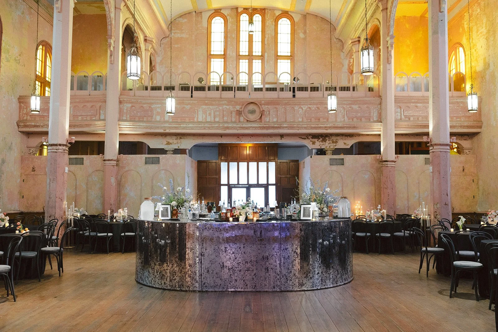 Interior of an elegant, vintage-style banquet hall with a central bar, surrounded by tables with black chairs, floral arrangements, and candles; high arched windows and hanging lanterns