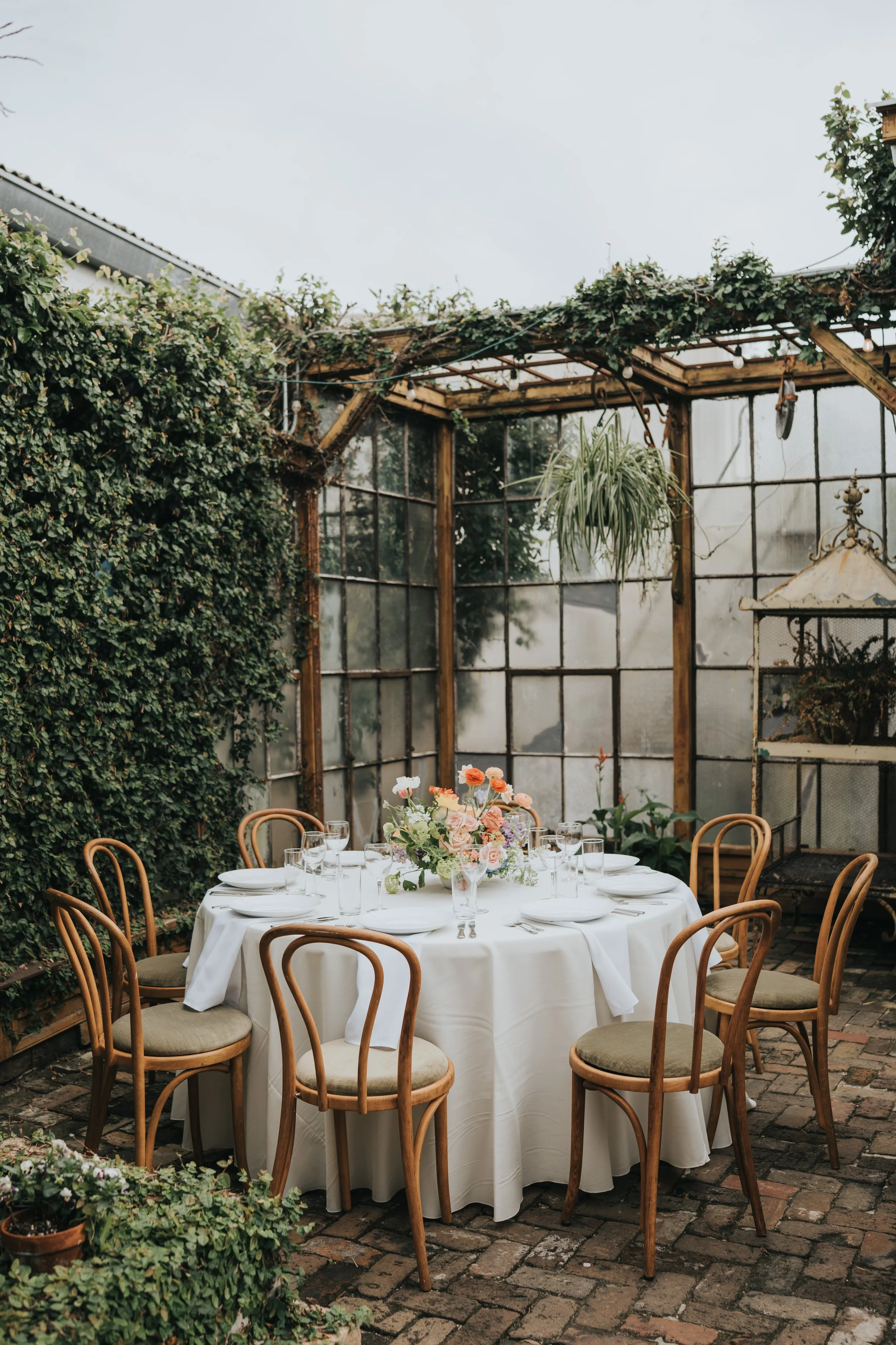 A round table with a white tablecloth set for a meal with wine glasses, plates, and a floral centerpiece in an outdoor patio with brick pavement, surrounded by green plants and a wooden greenhouse.