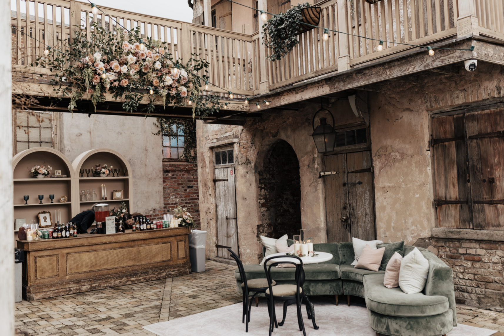 An indoor rustic venue decorated for a celebration, with a bar area, floral arrangements, a curved velvet sofa with pillows, and string lights hanging from the wooden balcony.