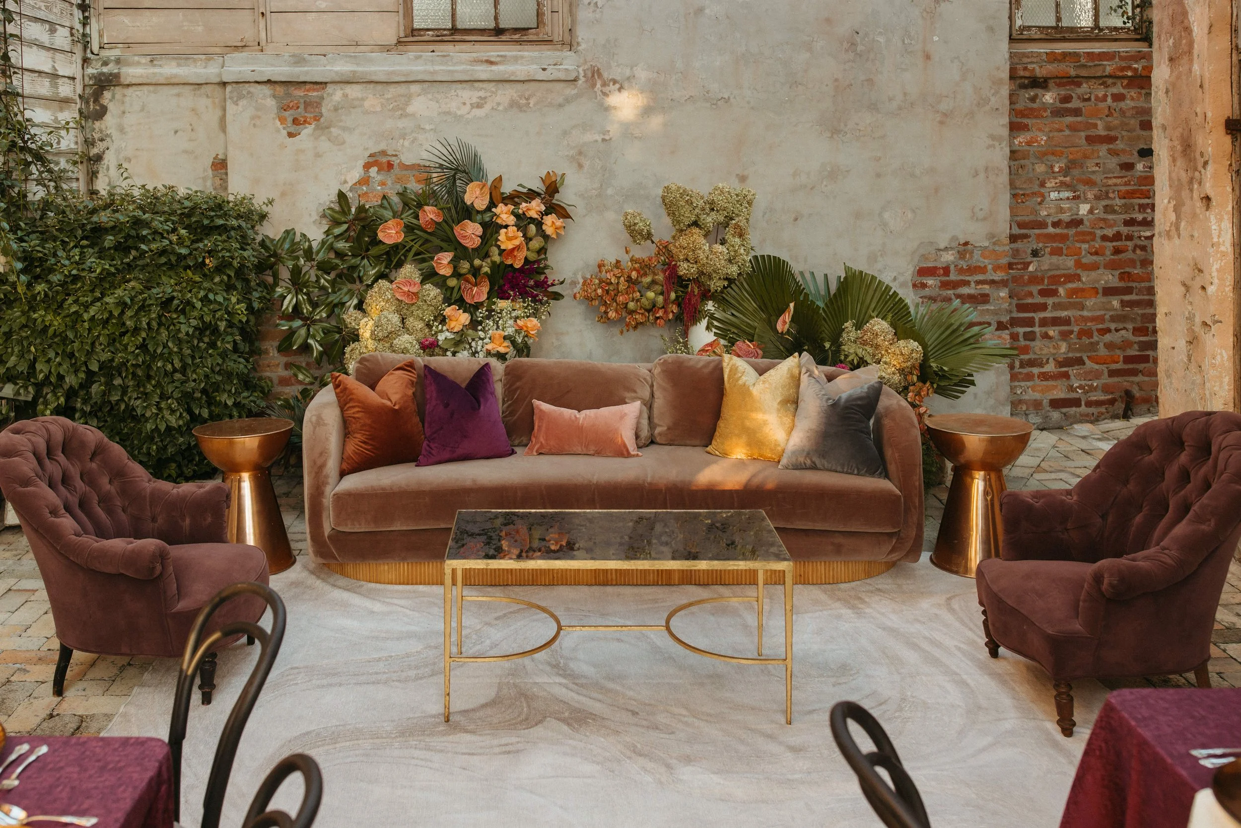 An indoor space with a rustic brick and plaster wall, decorated with colorful flowers and green foliage. There are two velvet armchairs on each side and a sofa in the center with various velvet cushions in gold, purple, pink, and gray. A gold coffee 