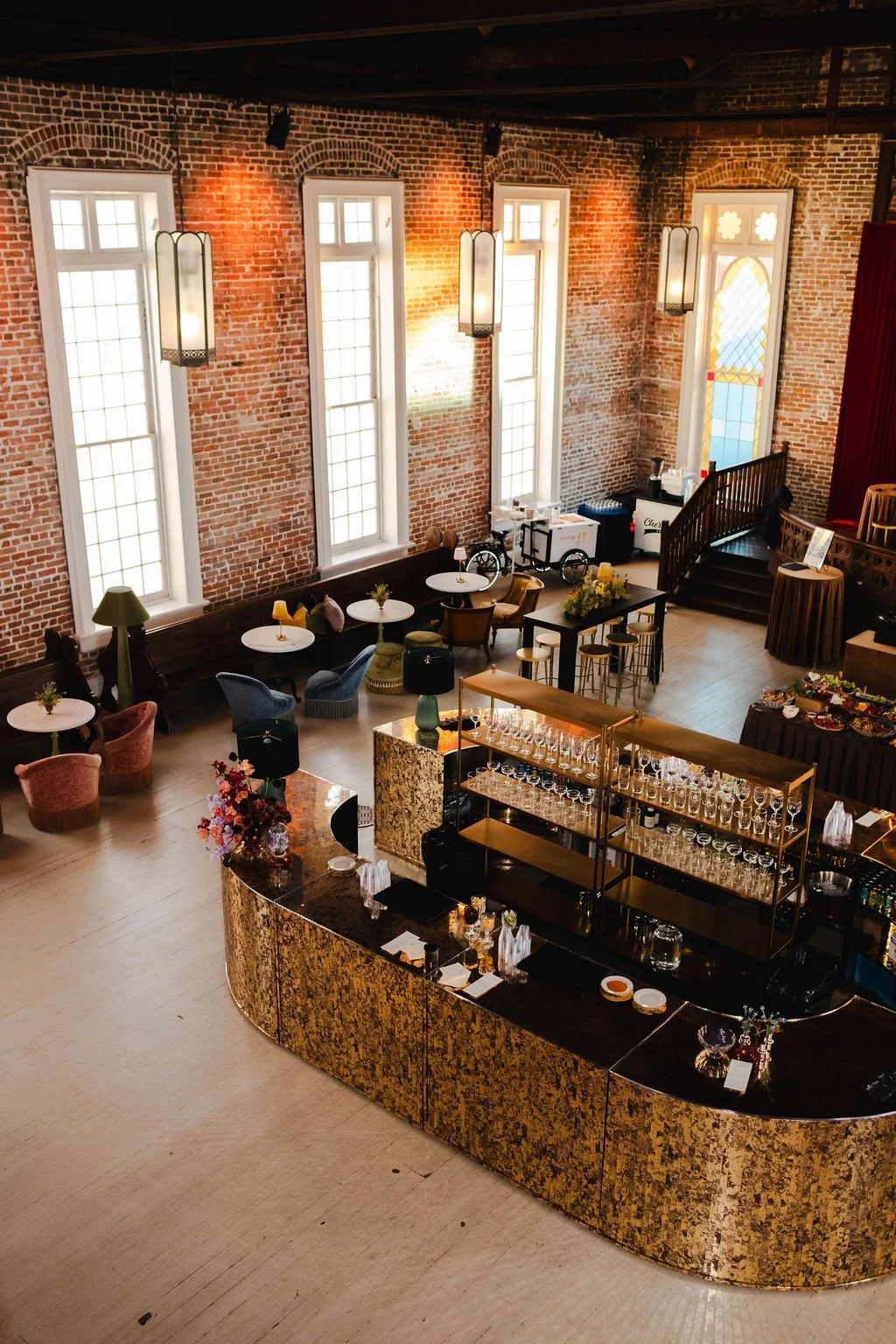 Interior of a stylish event space with exposed brick walls, tall stained-glass windows, and a curved gold and black bar counter with glassware. There are seating areas with chairs and small tables, a staircase near the back, and decorative lighting fixtures hanging from the ceiling.