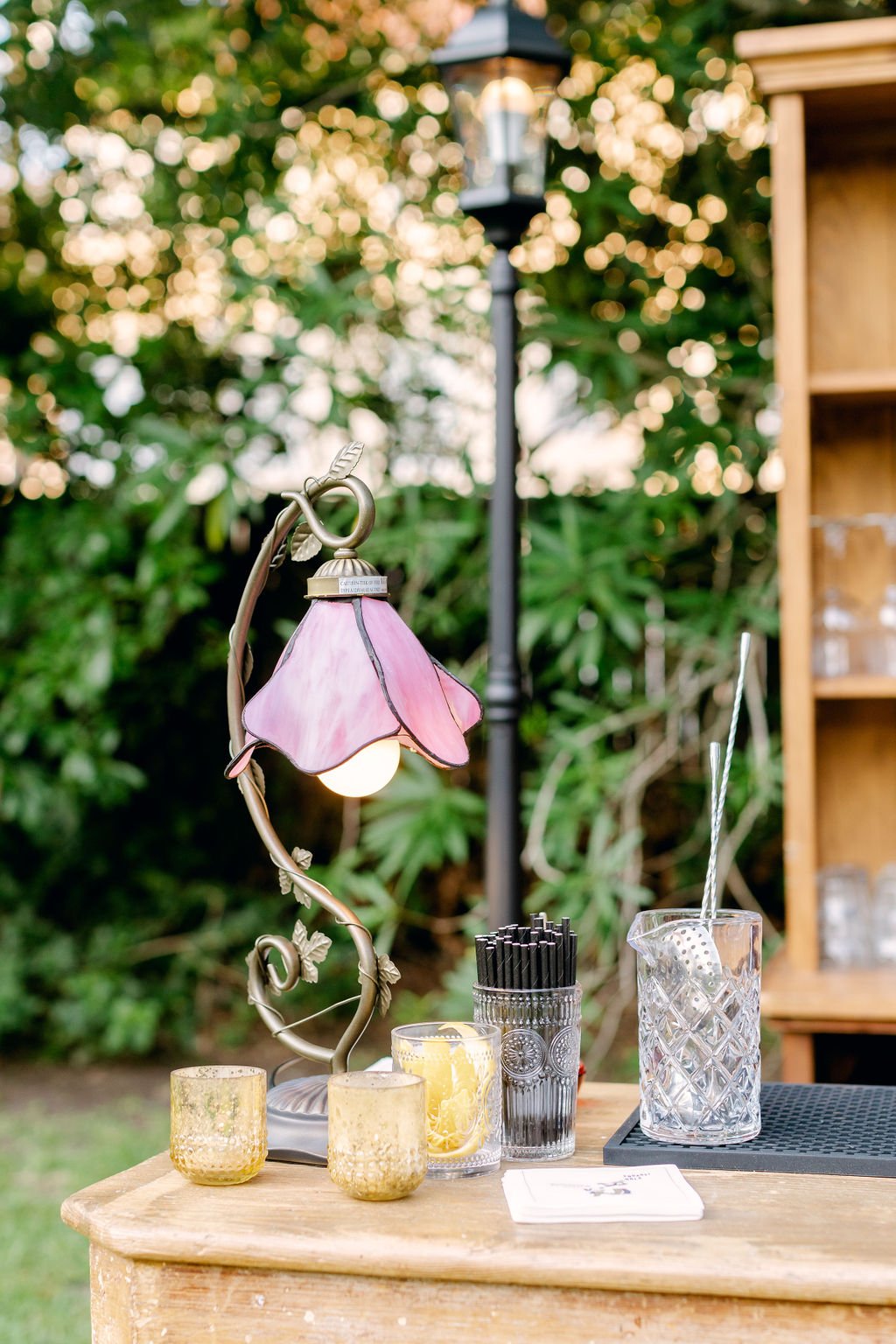 Outdoor setting with a decorative pink flower-shaped lamp, glass jars, and a clear glass with a straw on a wooden table, with lush greenery in the background.