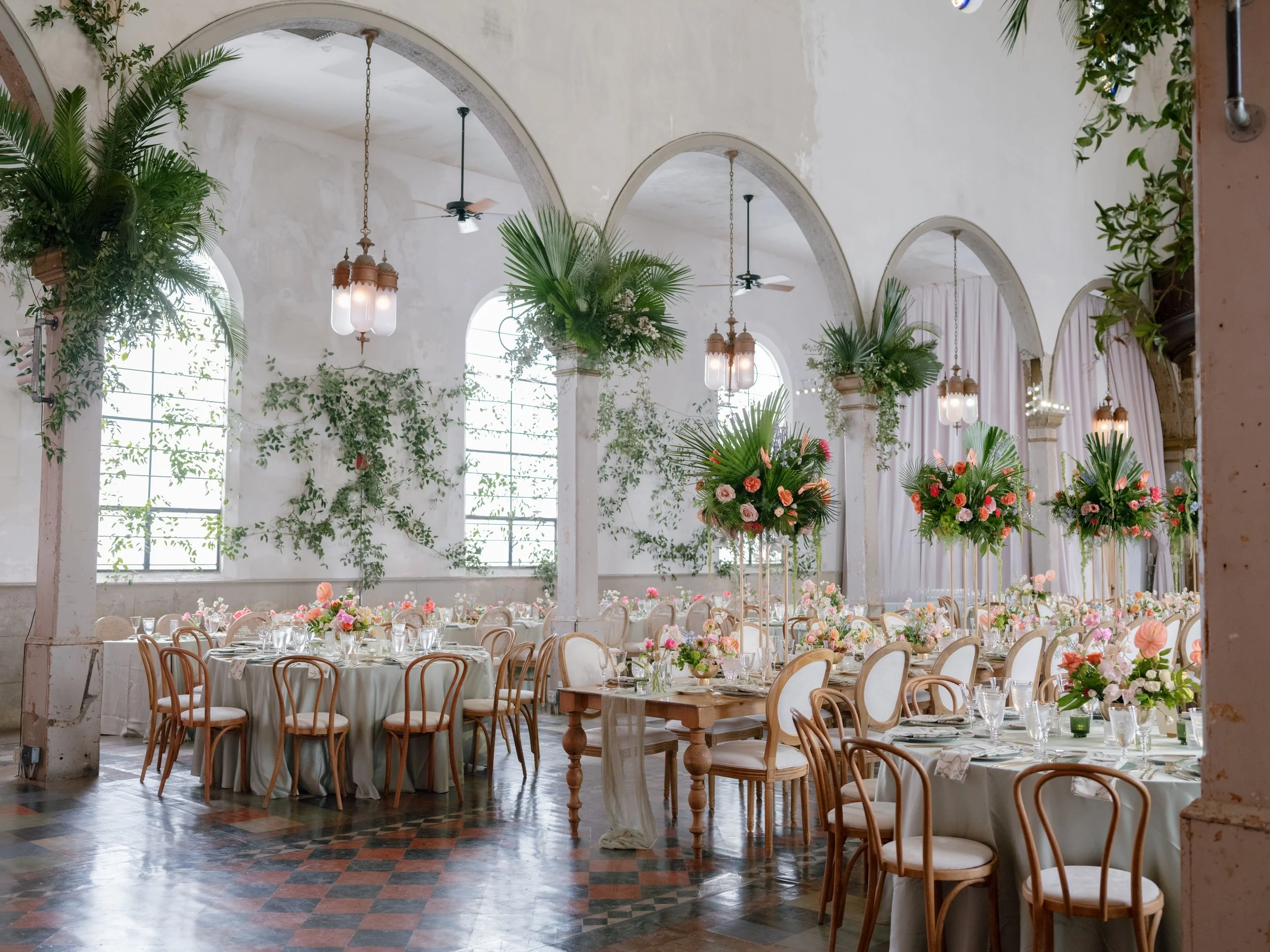 Elegant banquet hall decorated with lush green foliage and pink flowers, set for a formal event with round tables, white tablecloths, and wooden chairs. Large arched windows let in natural light, and white curtains hang along the walls. Hanging lante
