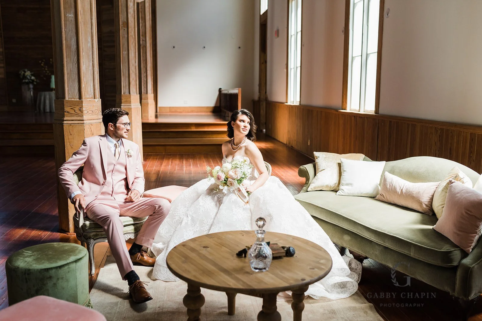 A bride and groom sitting in a room with wooden floors and paneling, large windows, a green sofa with pillows, and a round wooden coffee table with a glass decanter and small decorative items.