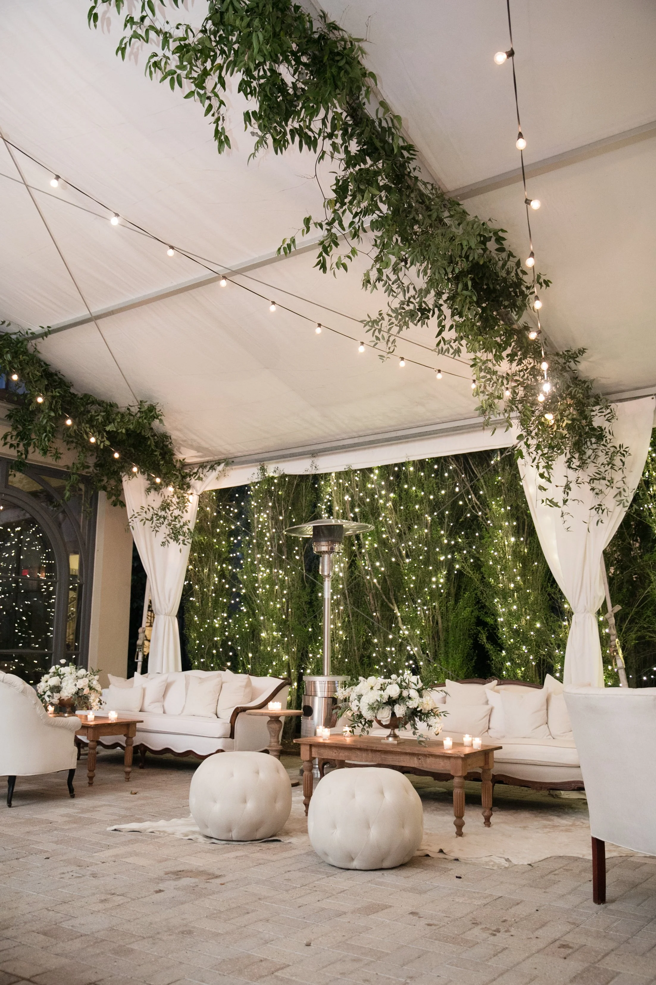 Elegant outdoor event space decorated with string lights, white curtains, floral arrangements, and white cushioned sofas and ottomans.