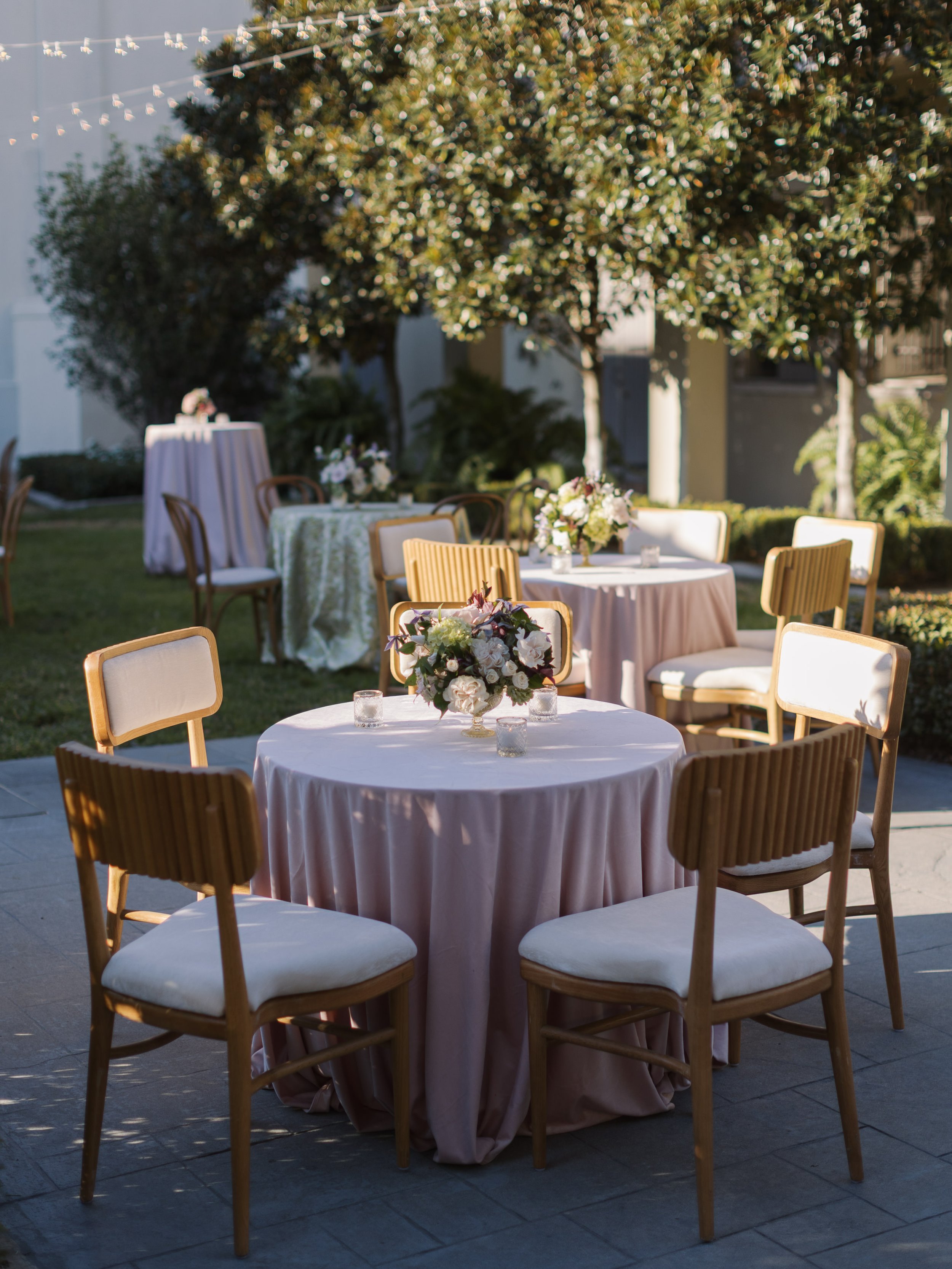 Outdoor wedding reception area with round tables draped in blush pink tablecloths and floral centerpieces. Wooden chairs with cream cushions surround the tables. Tall floral arrangement in the background, string lights hanging overhead, and a large t