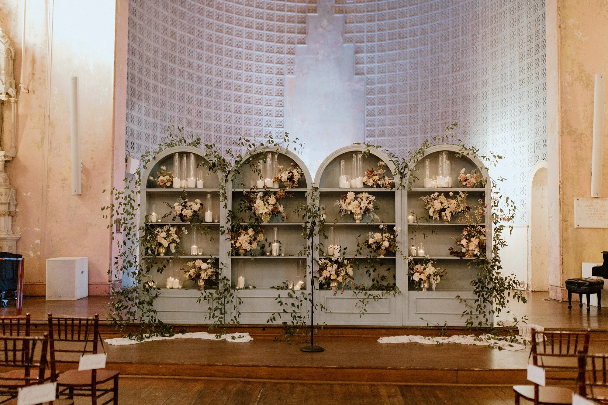 Wedding ceremony setup with floral arrangements, candles, and a white backdrop on a stage in a church or similar venue.