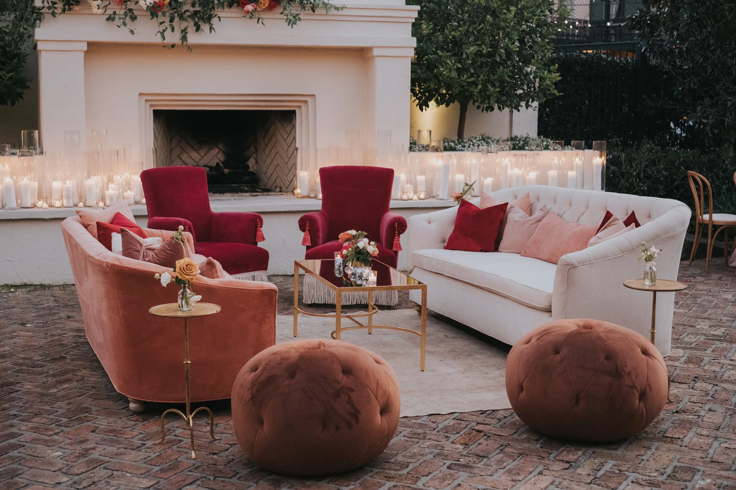 Outdoor seating area with modern sofas, velvet chairs, small tables, and candles around a fireplace adorned with flowers and greenery.