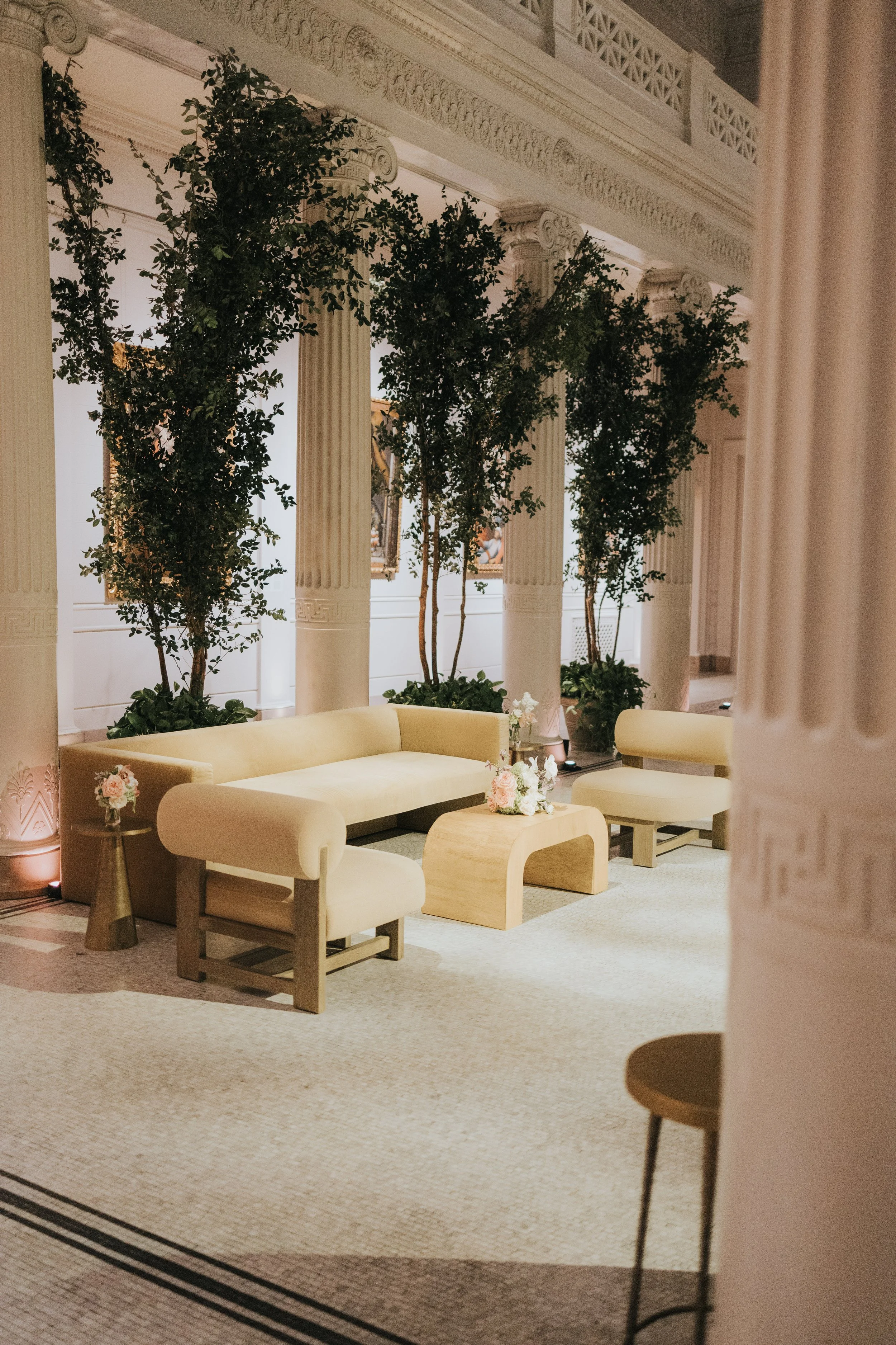 Elegant indoor space featuring cream-colored curved sofas, wooden tables with floral arrangements, tall potted trees, classical columns, and ornate crown molding in the background.
