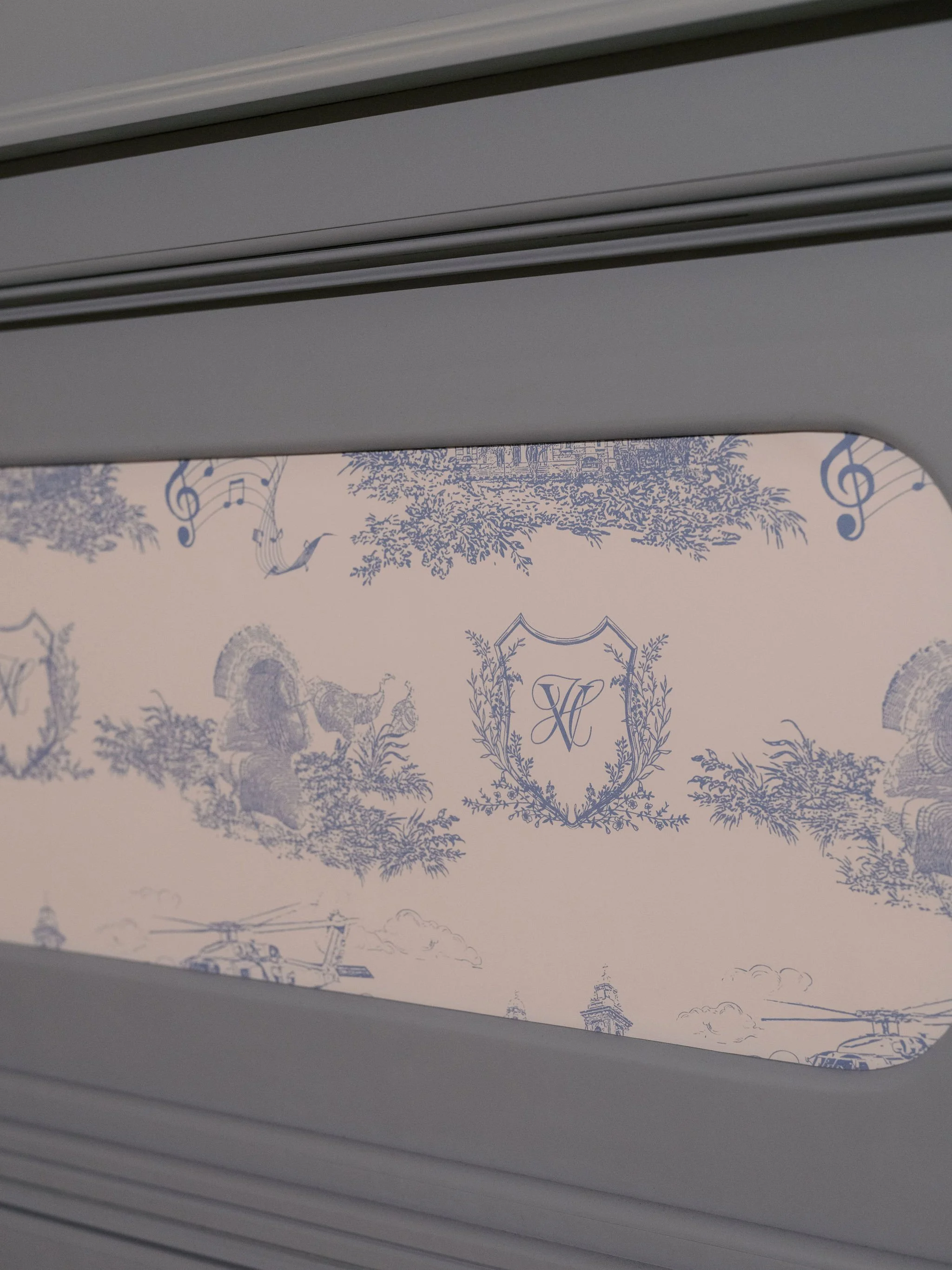 View of a decorative wall with blue illustrations of trees, musical notes, and heraldic symbols, seen through a horizontal slatted window or vent.