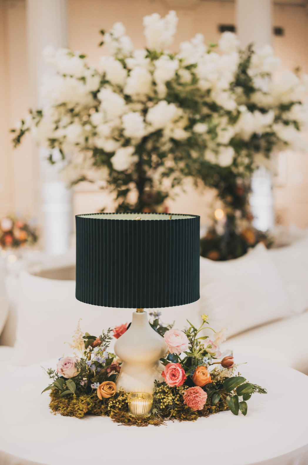 A table centerpiece with a white lamp and a floral arrangement of pink, orange, and purple flowers, surrounded by moss and a small candle, in front of a background of large white floral decoration.