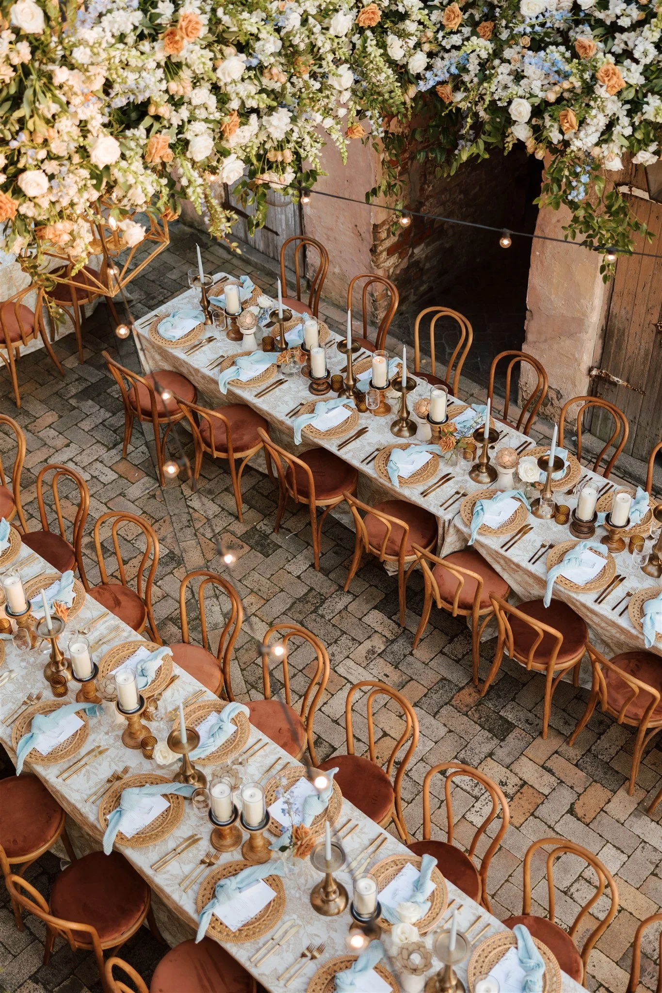 A long dining table set for an event, decorated with candles, floral centerpieces, and place settings with plates, napkins, and cutlery. Overhead, a large arrangement of flowers hangs from the ceiling, and string lights add a warm ambiance. The setti