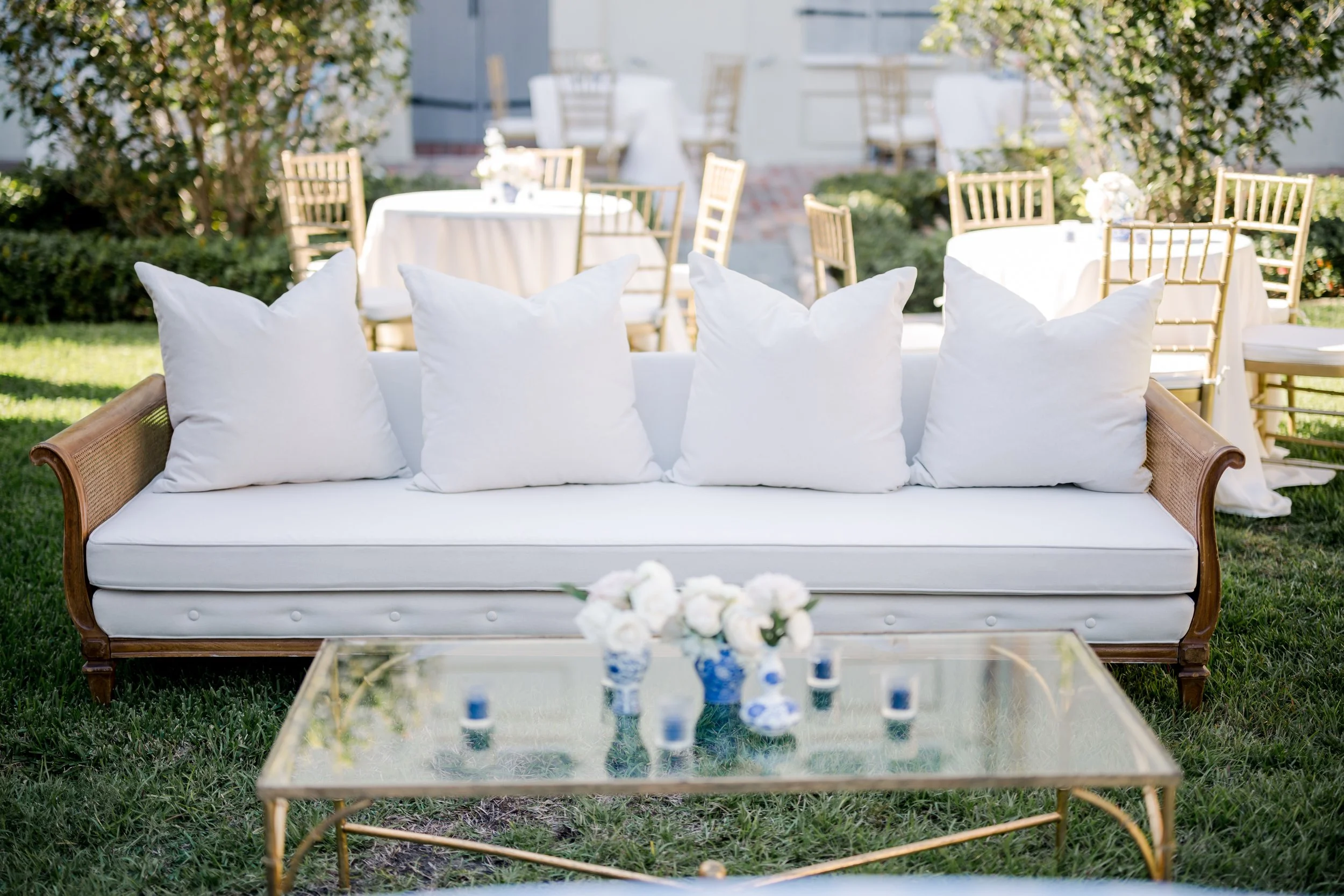 Outdoor patio with a white sofa with four white pillows, a glass coffee table with a floral centerpiece and small bottles, in front of several round tables with white tablecloths and gold chairs, surrounded by greenery.